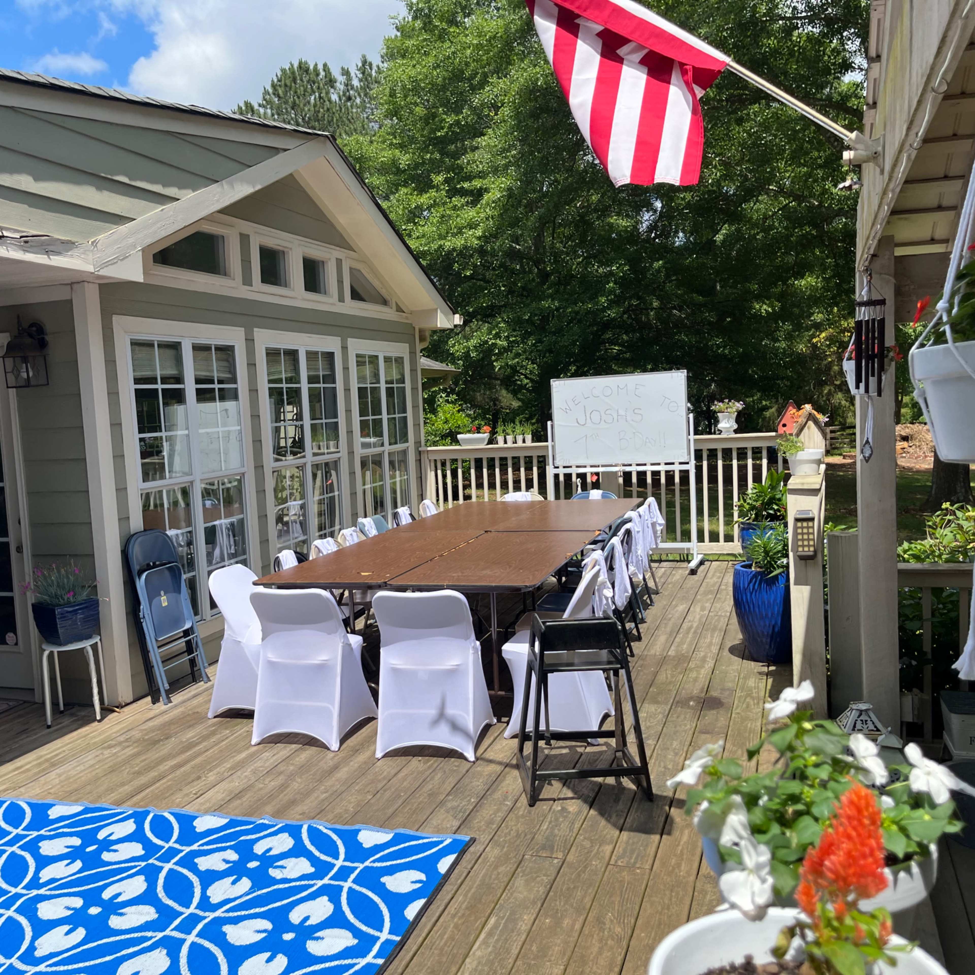 A spacious deck features a long table set with white chairs, surrounded by greenery and decorative plants, with a flag and a whiteboard in the background.