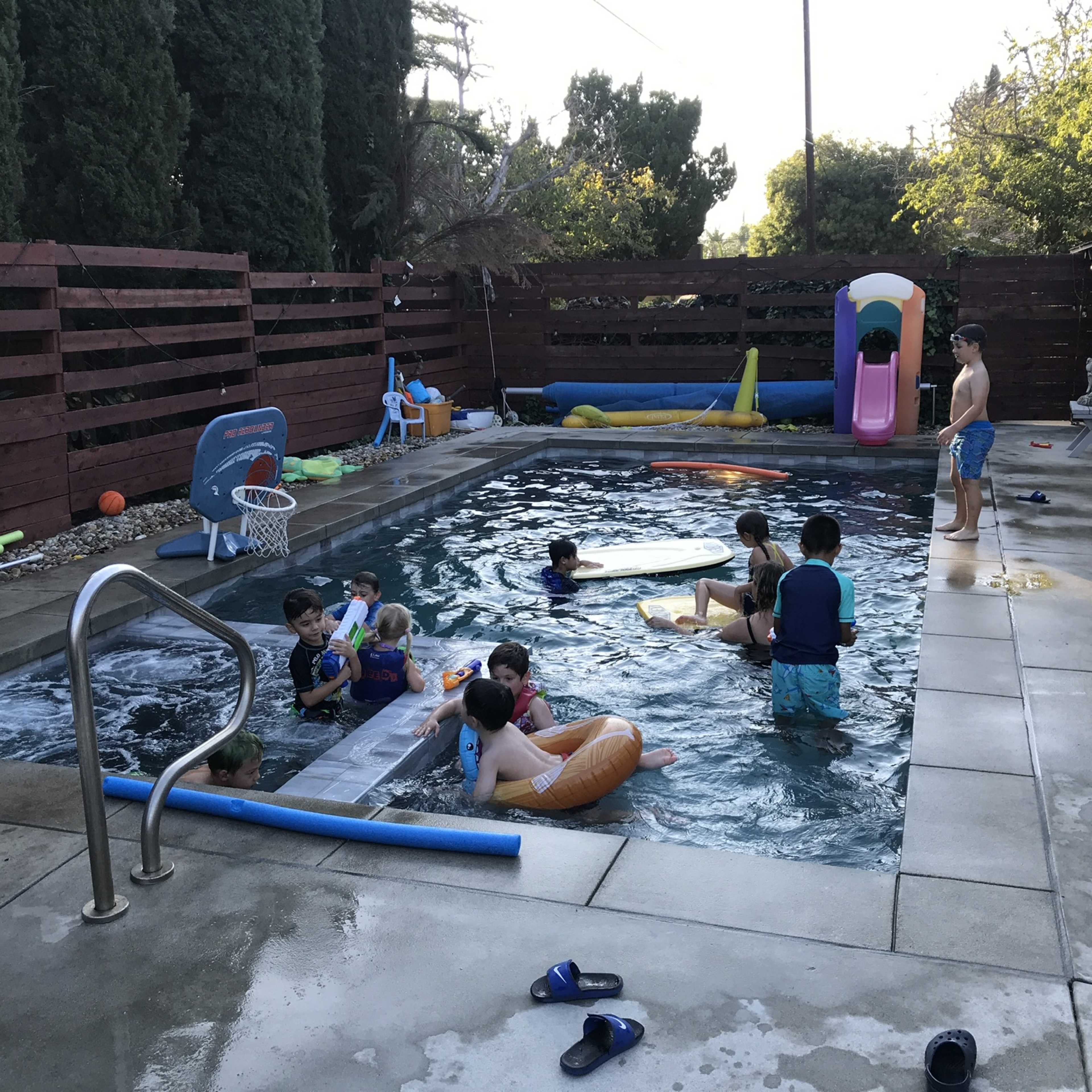 A group of children is playing in a swimming pool surrounded by a patio area with toys and inflatable floaters.
