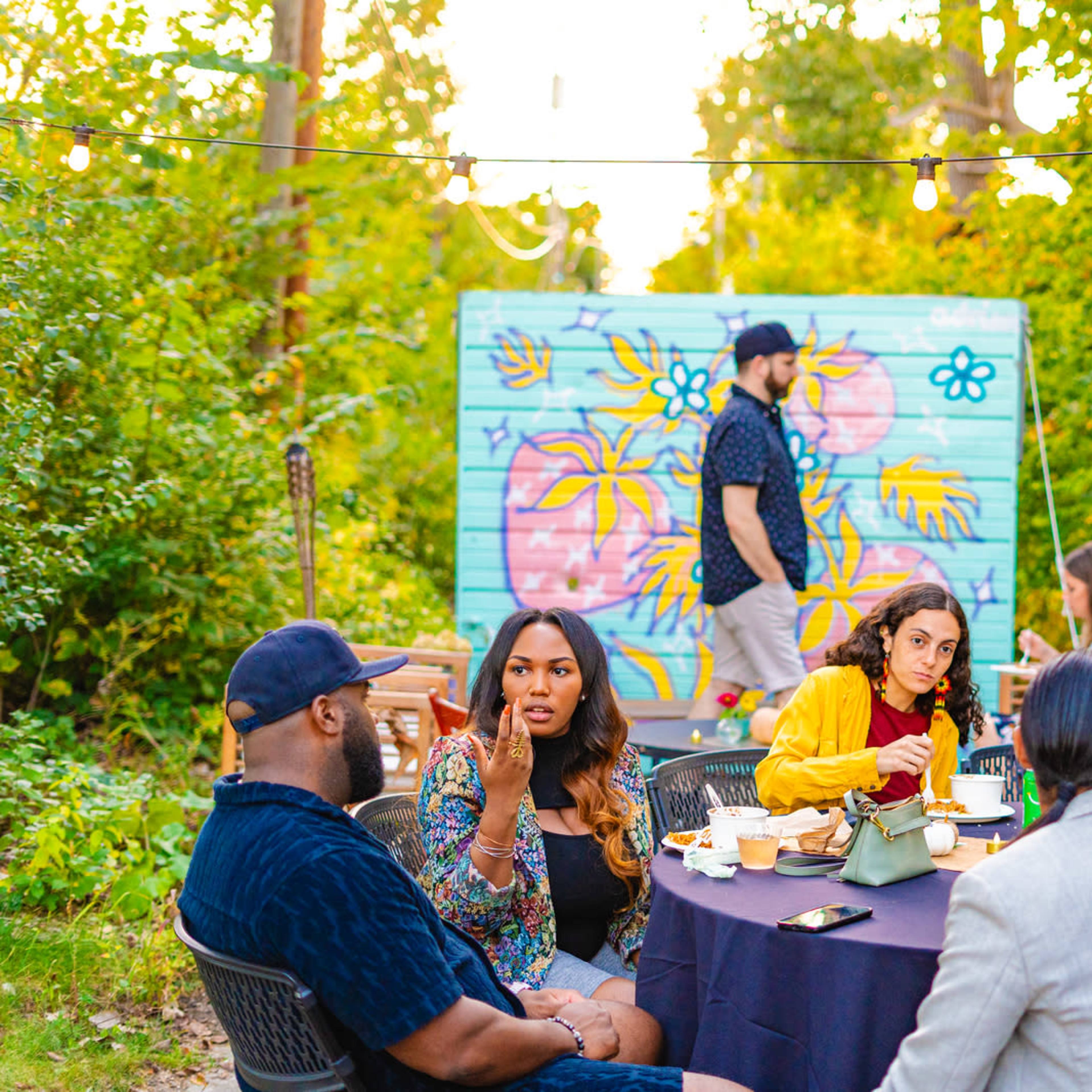 A group of people is seated around a table enjoying a meal in a garden setting with a colorful mural in the background.