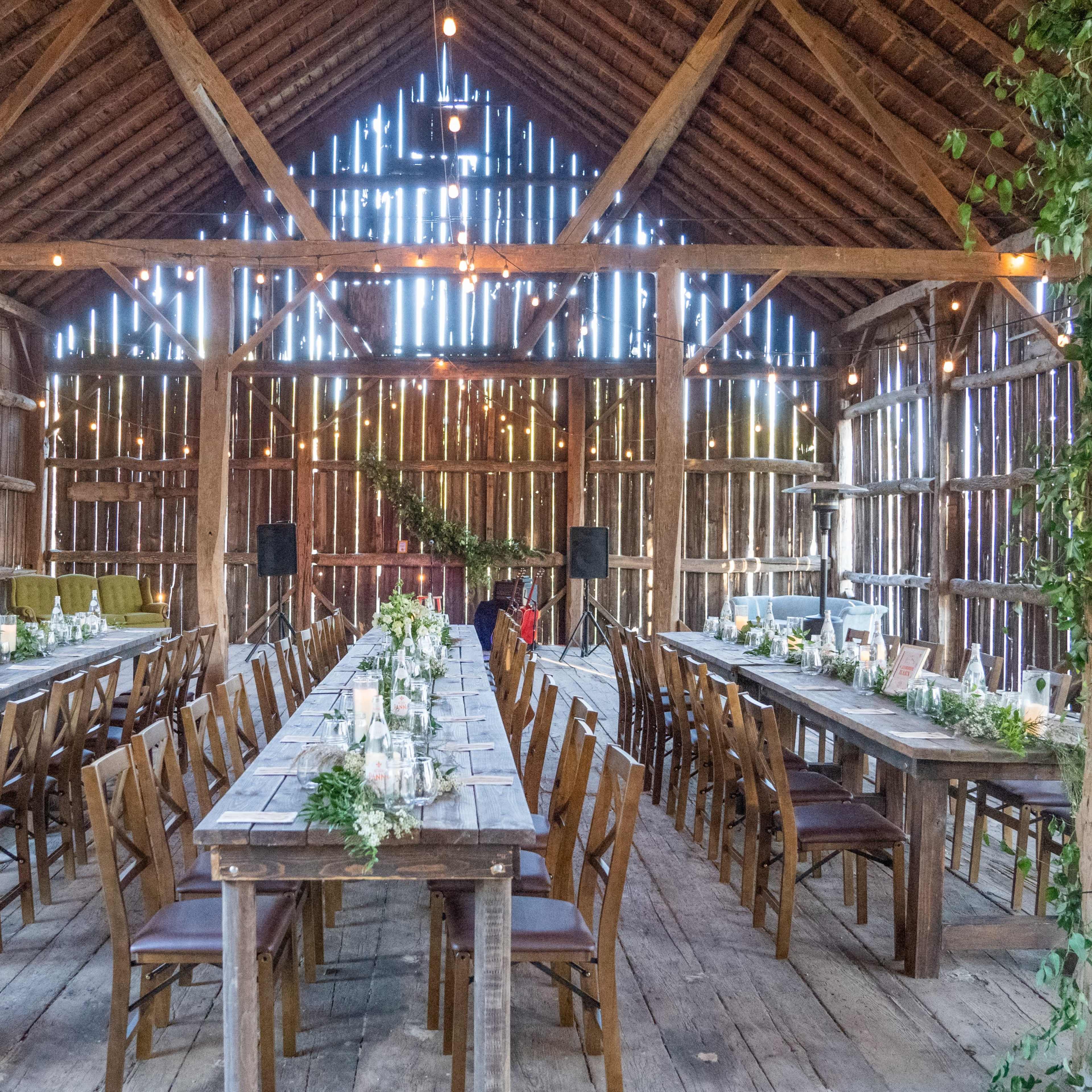 The interior of a barn features two long wooden tables set with candles and floral arrangements, flanked by chairs, under a ceiling adorned with string lights.