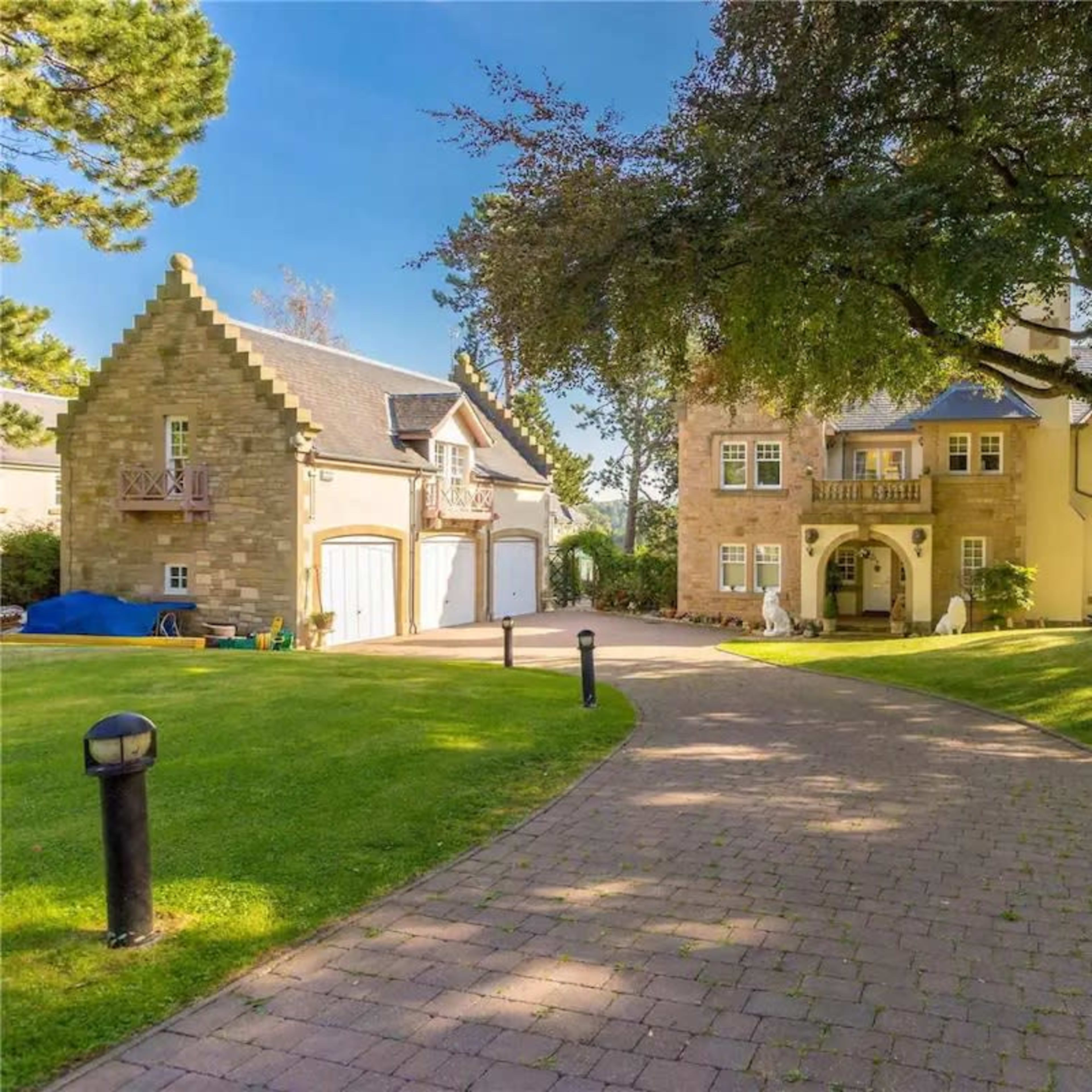 A large house with a tower is set alongside a two-car garage, surrounded by manicured lawns and trees.
