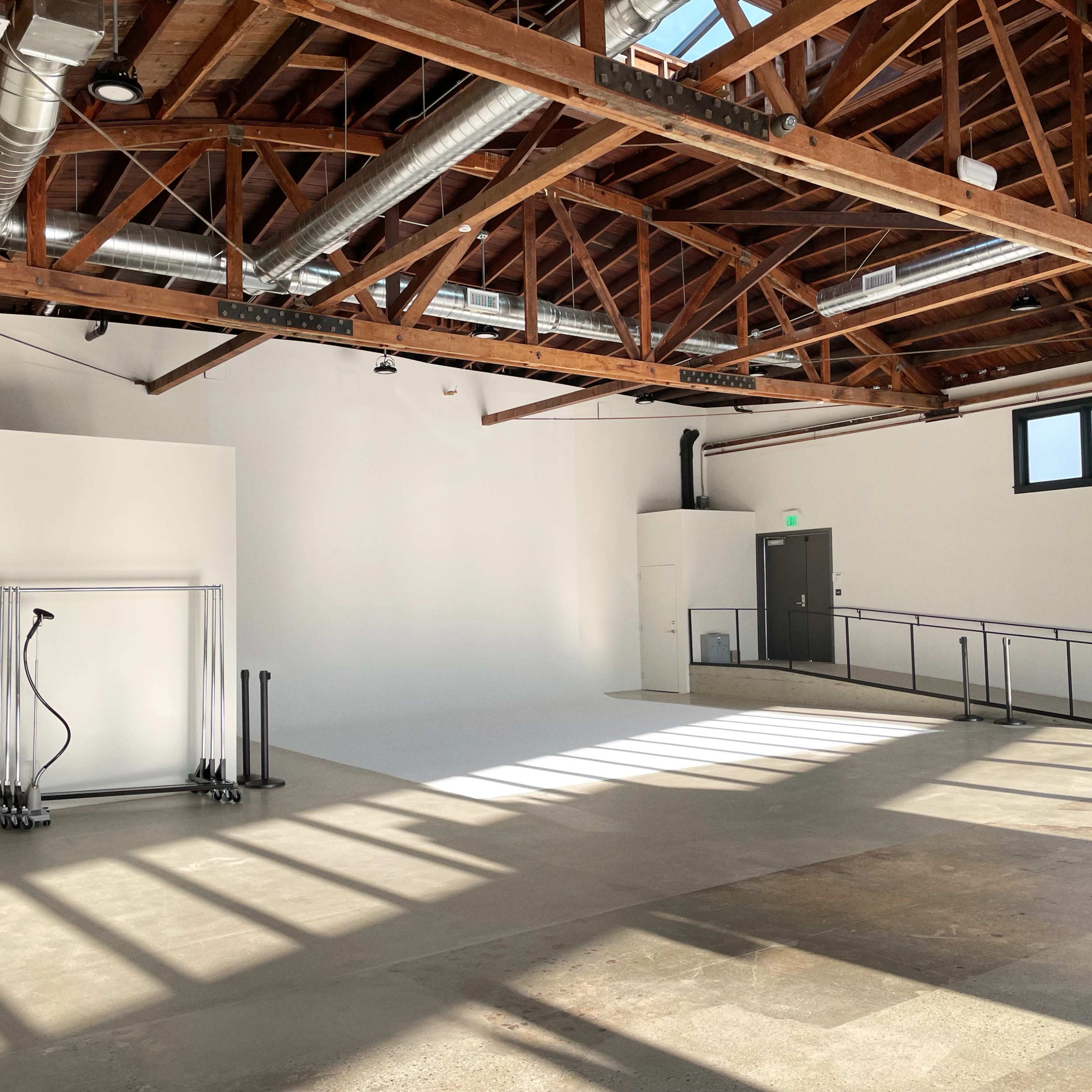A spacious interior with high wooden ceilings, a blank white wall, and a gray floor, illuminated by natural light streaming through large windows.