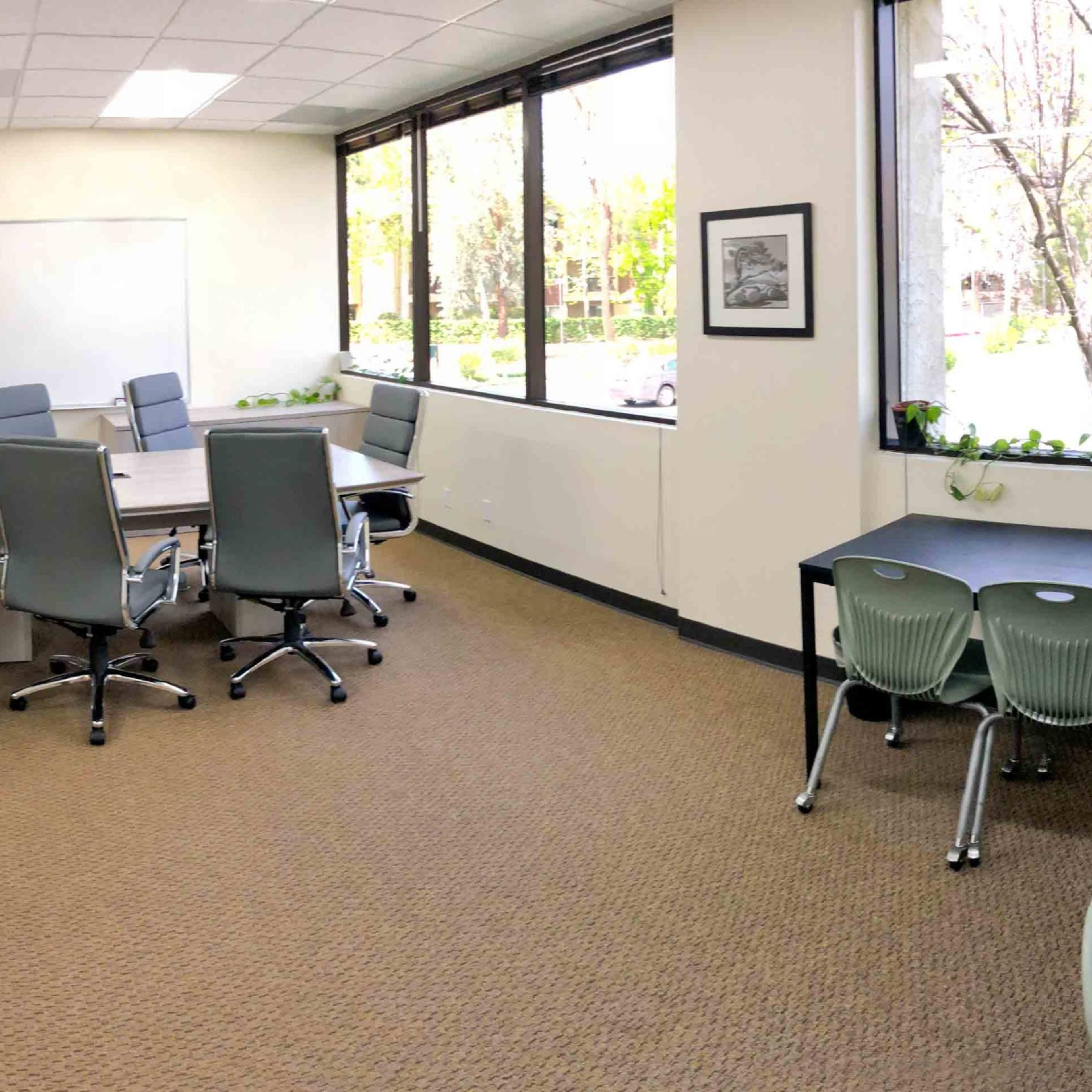 A modern classroom features a long, conference-style table surrounded by ergonomic chairs, with large windows providing natural light.