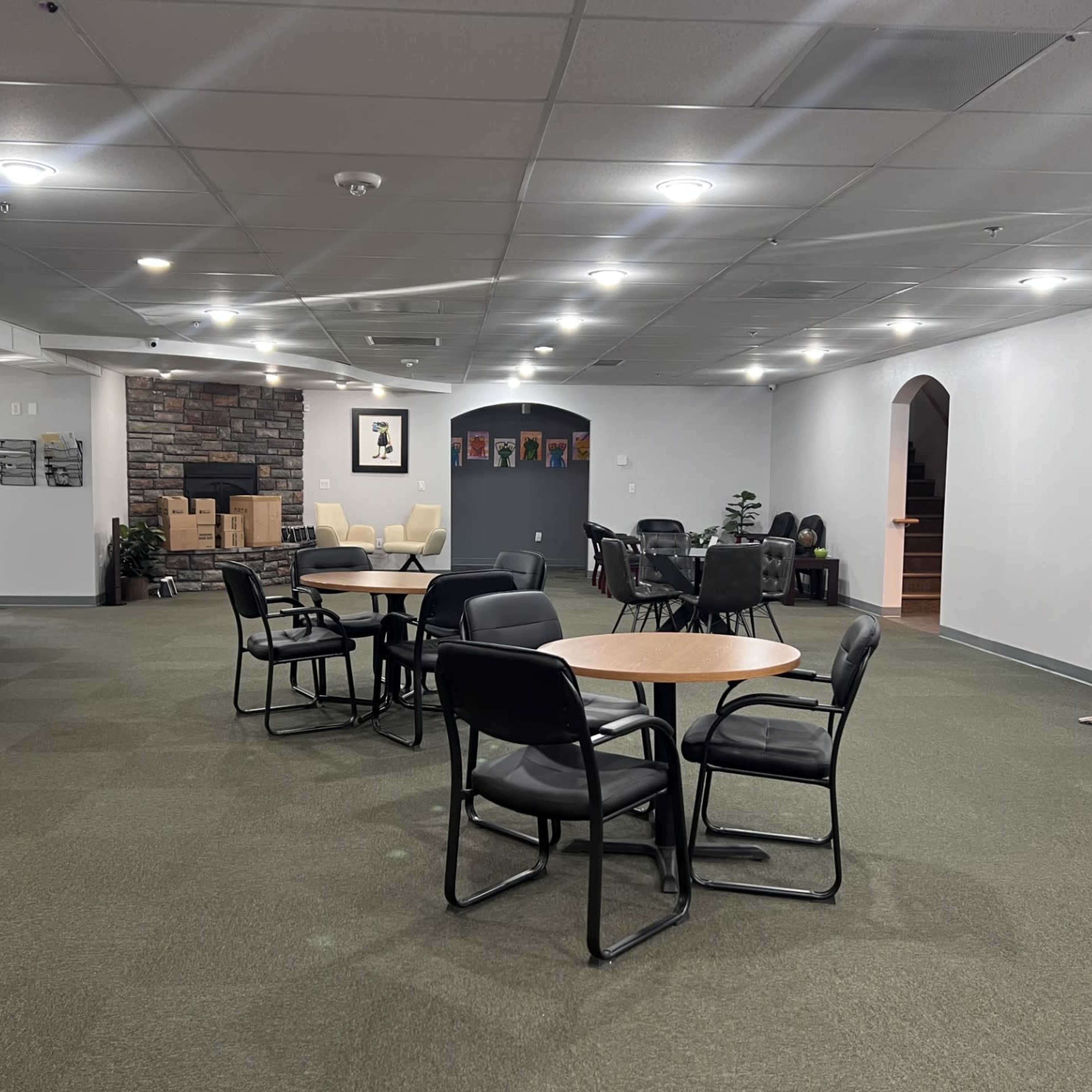 A spacious indoor area with several round tables and black chairs, featuring a varied seating arrangement, a stone wall, and an opening leading to another room.