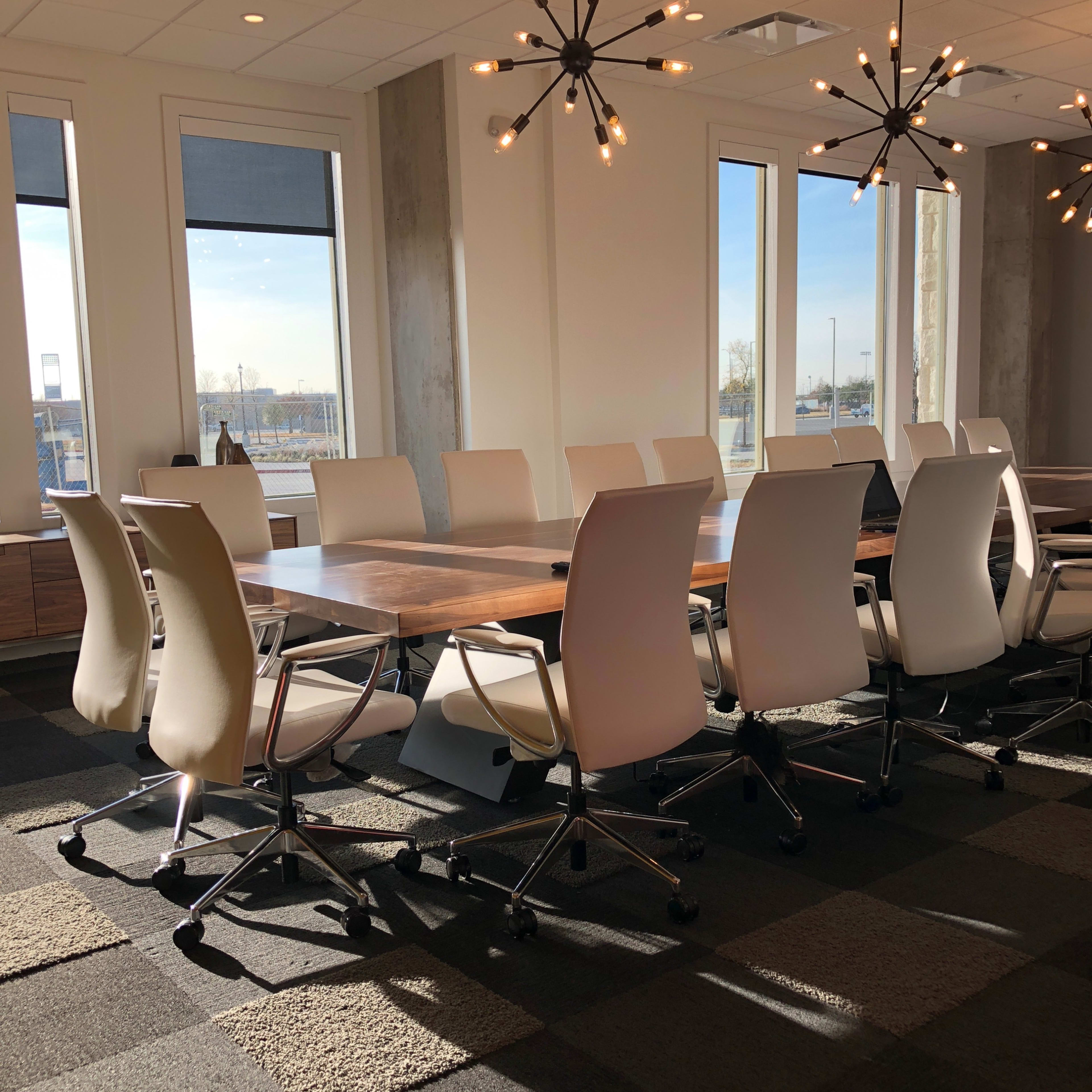 A conference room features a large table surrounded by ergonomic chairs and modern light fixtures, with windows providing natural light.
