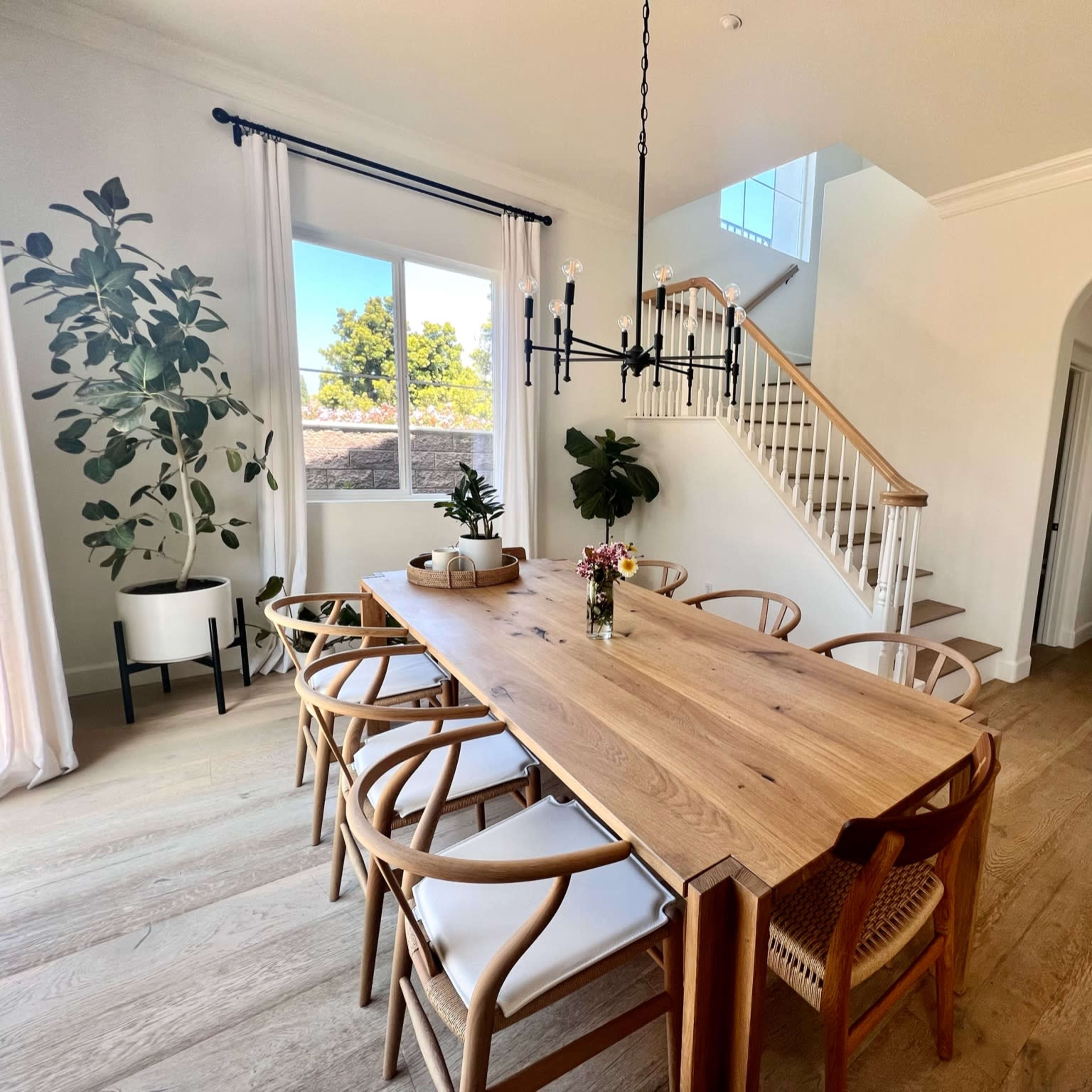 A dining area features a wooden table surrounded by chairs, with large windows and a staircase visible in the background.