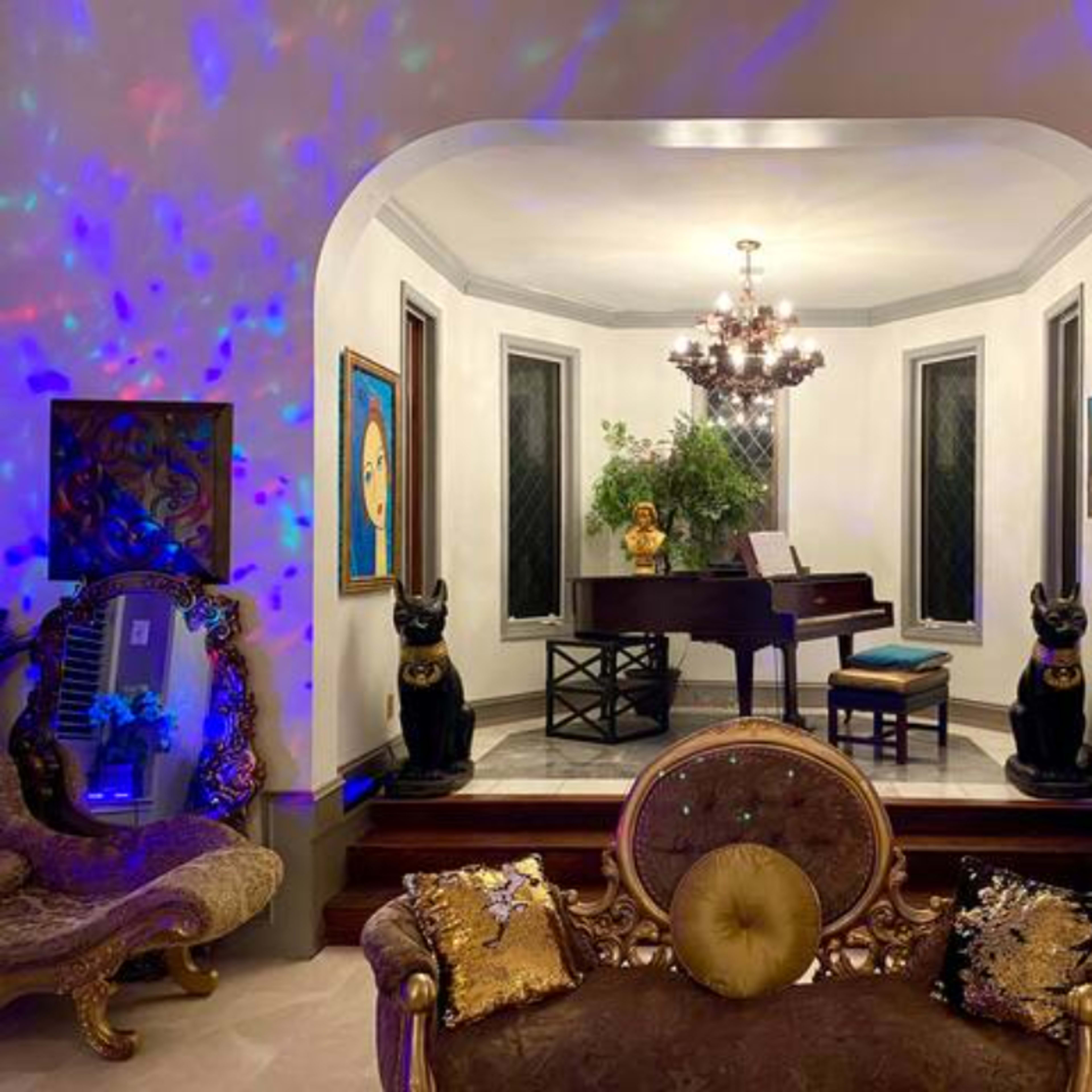 A grand room features a piano in the center, flanked by two ornate black statues, with colorful lights reflecting on the walls and various artworks displayed.