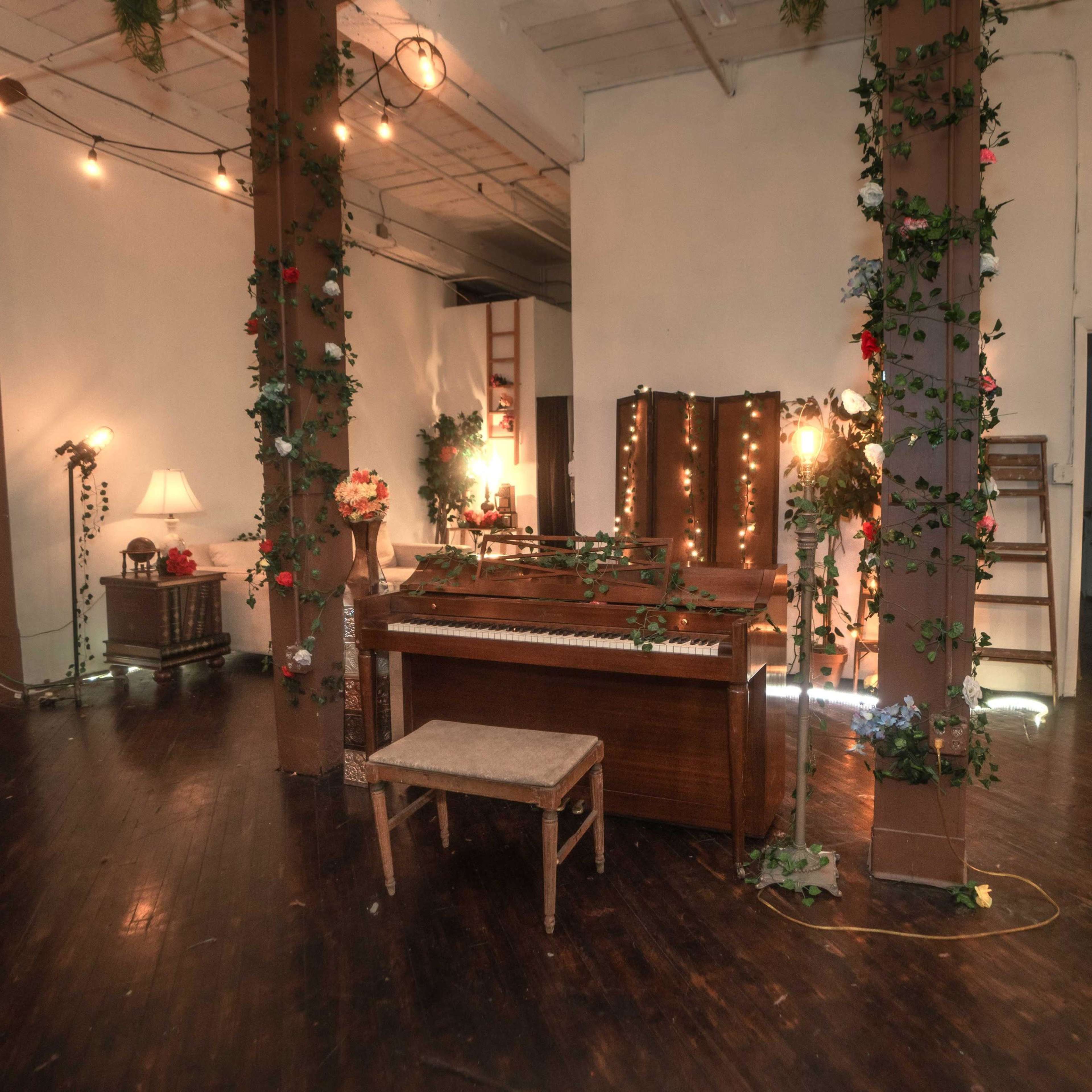 A spacious room with wooden floors, featuring a vintage piano surrounded by decorative plants and soft lighting from several lamps.