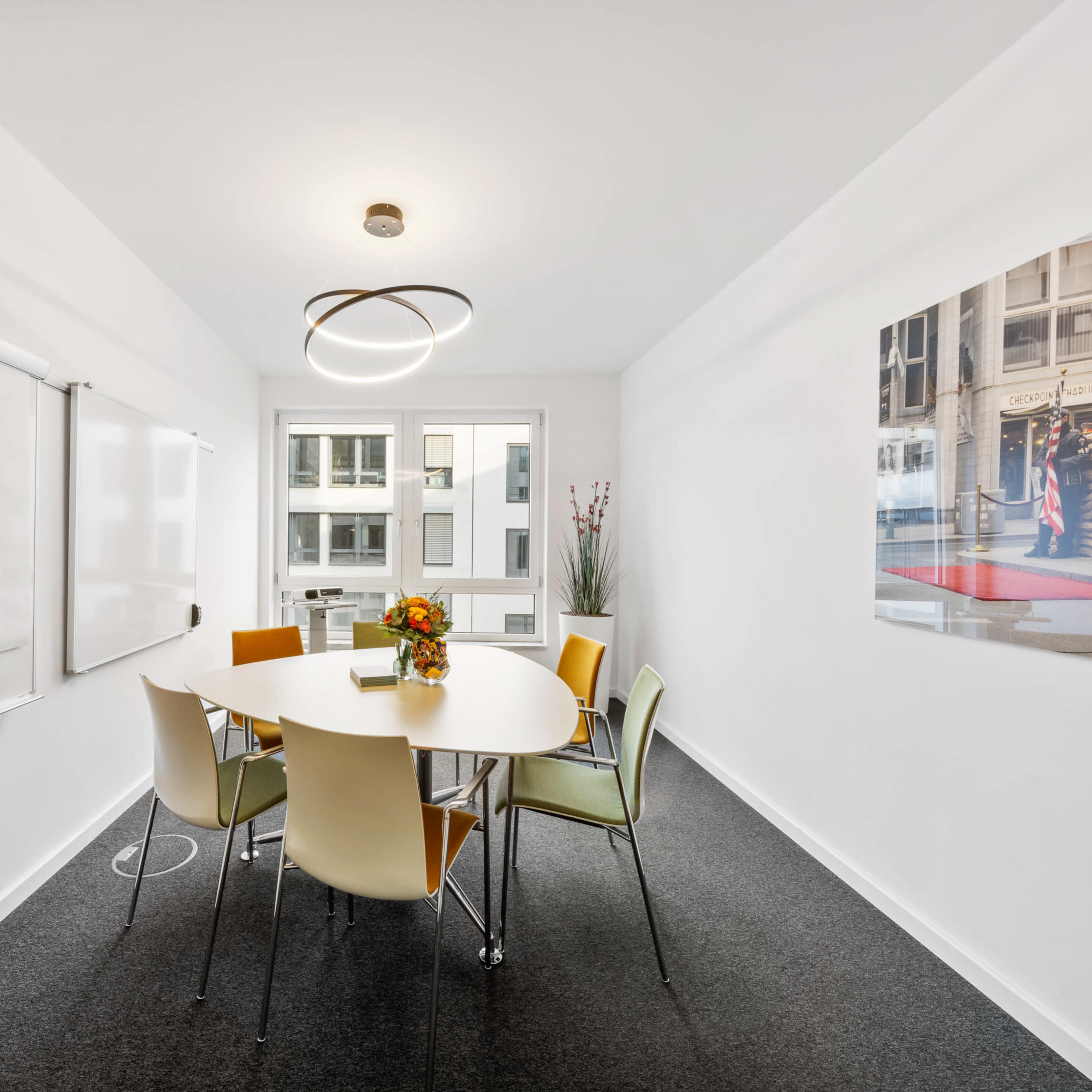 A modern conference room features a round table surrounded by six chairs, with a whiteboard and a window allowing natural light.