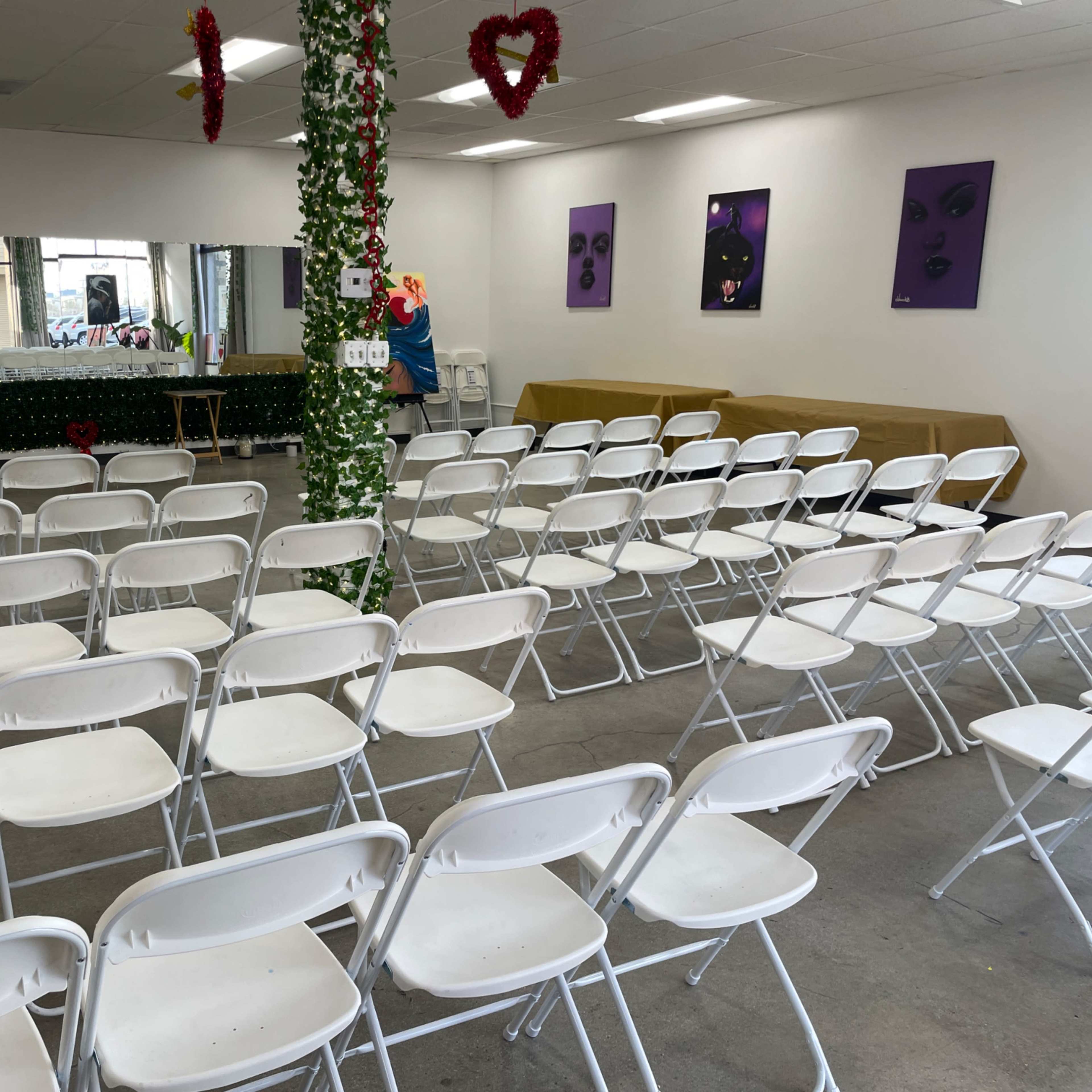 A row of white folding chairs arranged in a clear space, with a mirror and purple artwork on the walls.