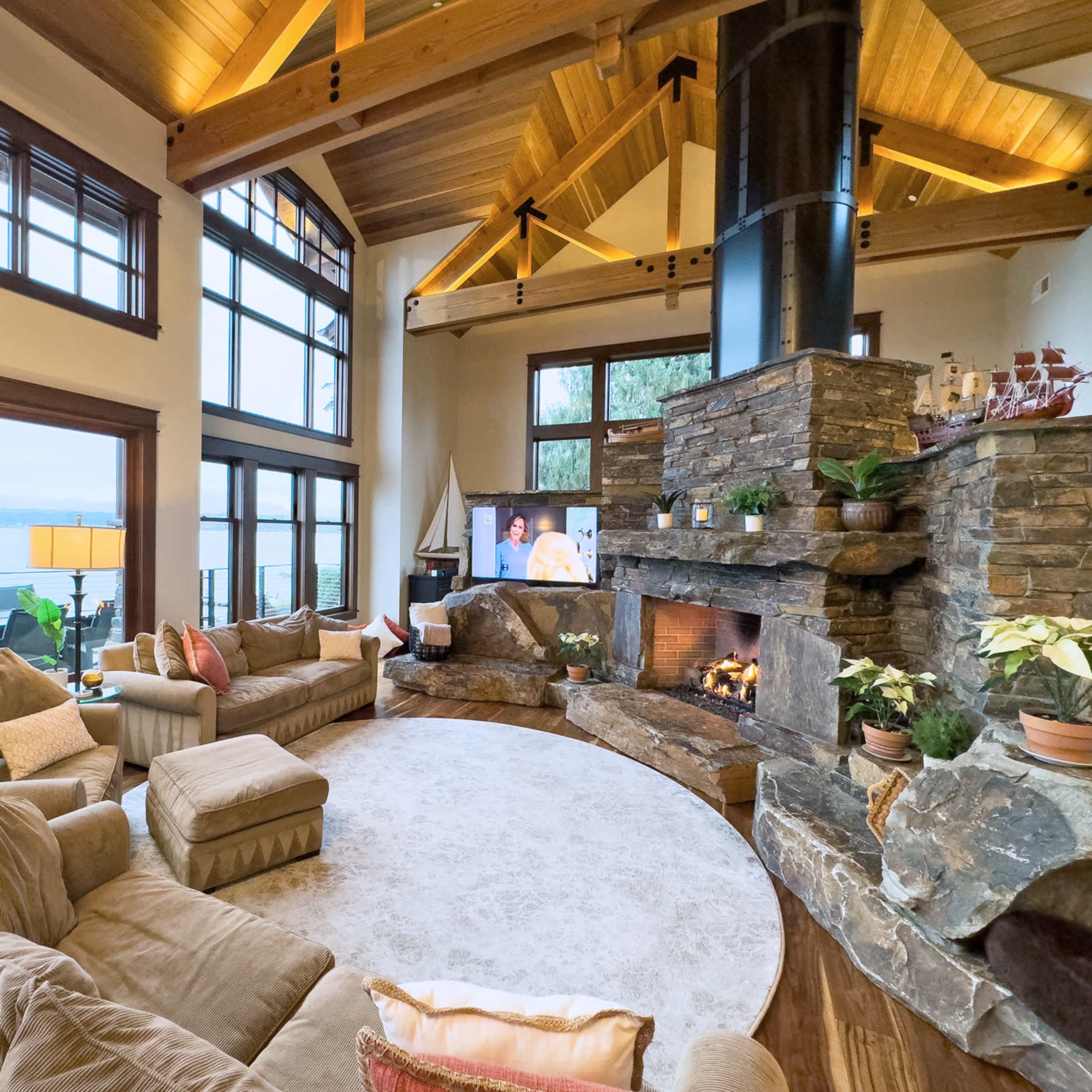 The spacious living room features a circular seating arrangement around a stone fireplace, with large windows providing a view of the lake outside.