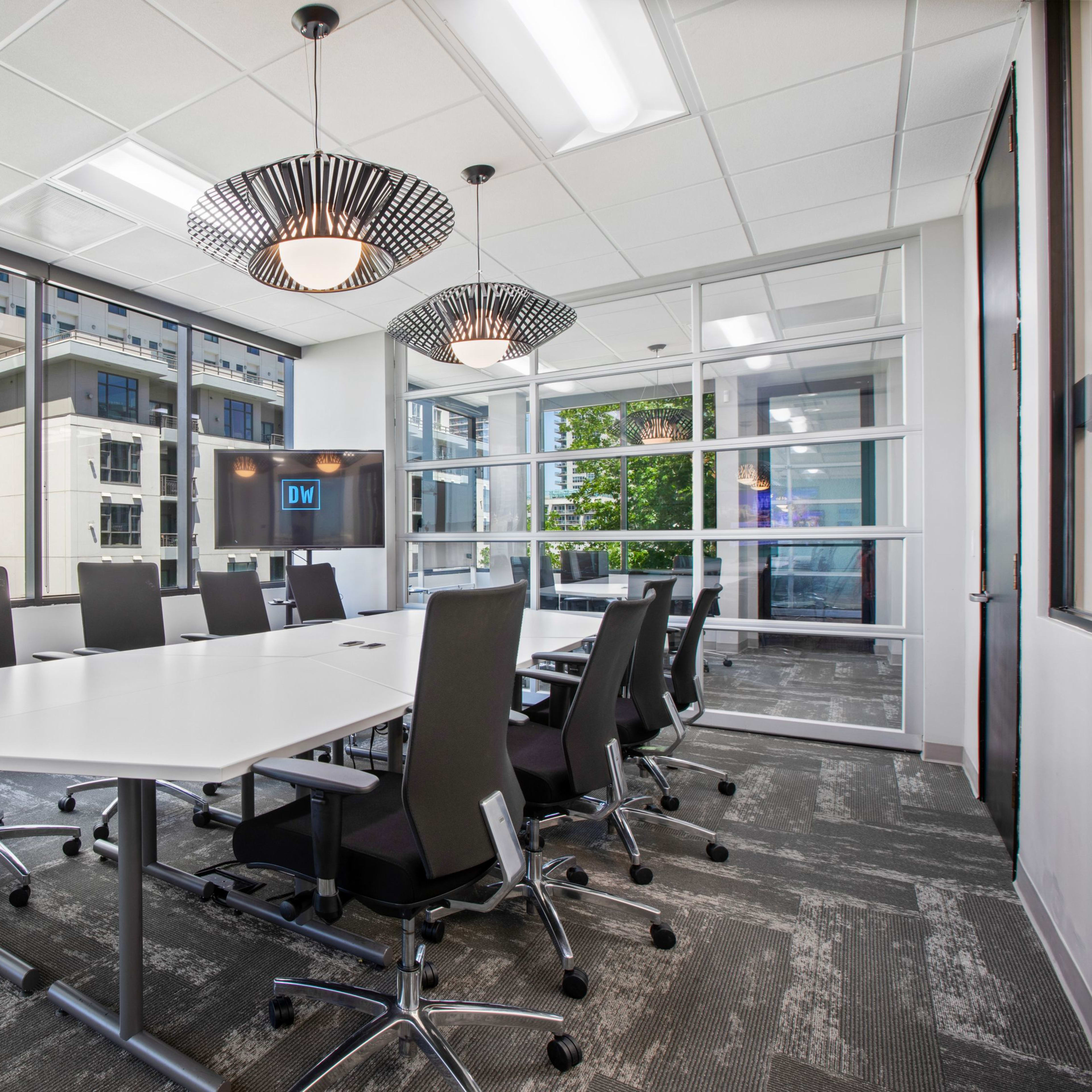 A modern conference room features a large white table surrounded by black chair, with windows providing natural light and a view of the outside.