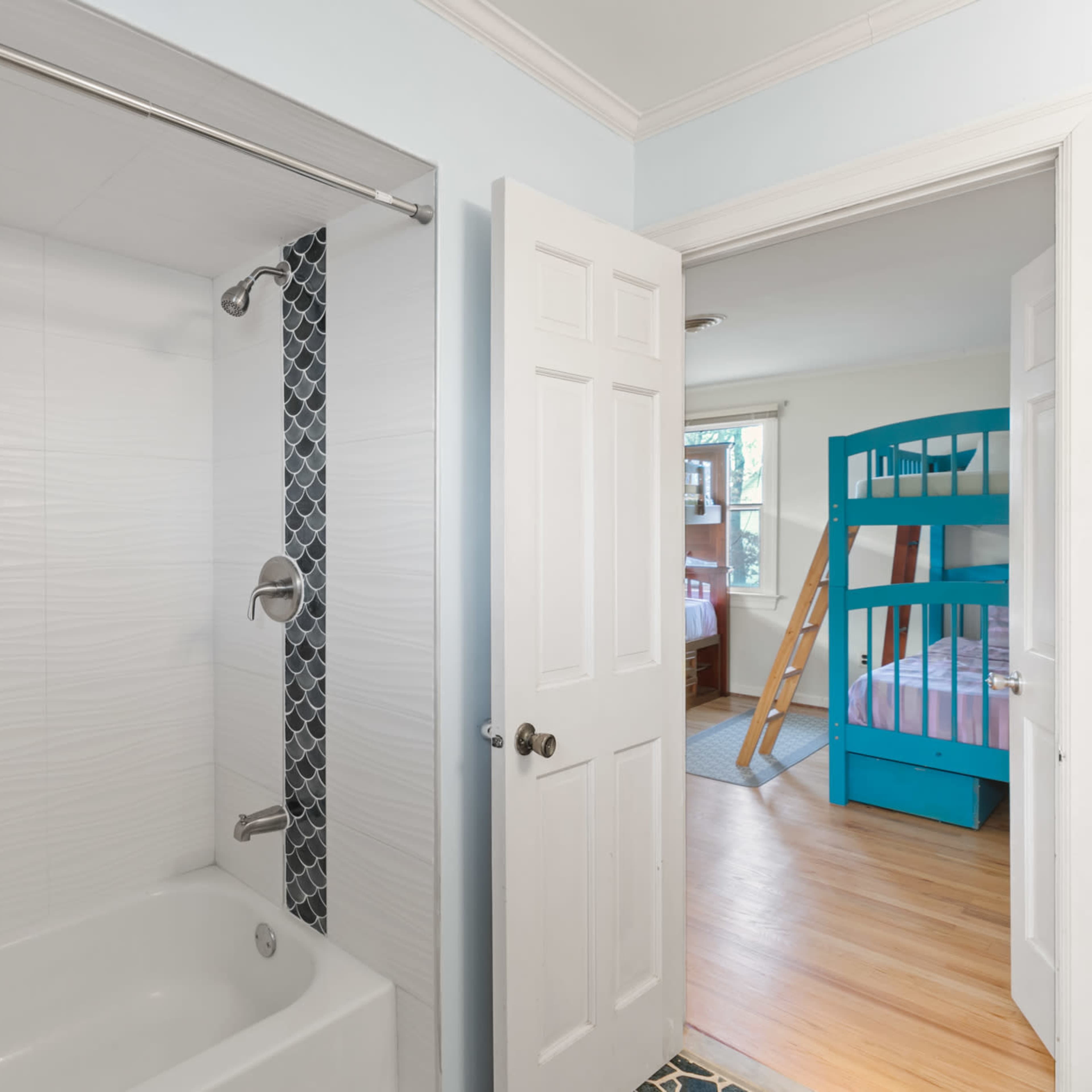A bathroom with a shower area and two open doors leading to a well-lit bedroom featuring a bunk bed.
