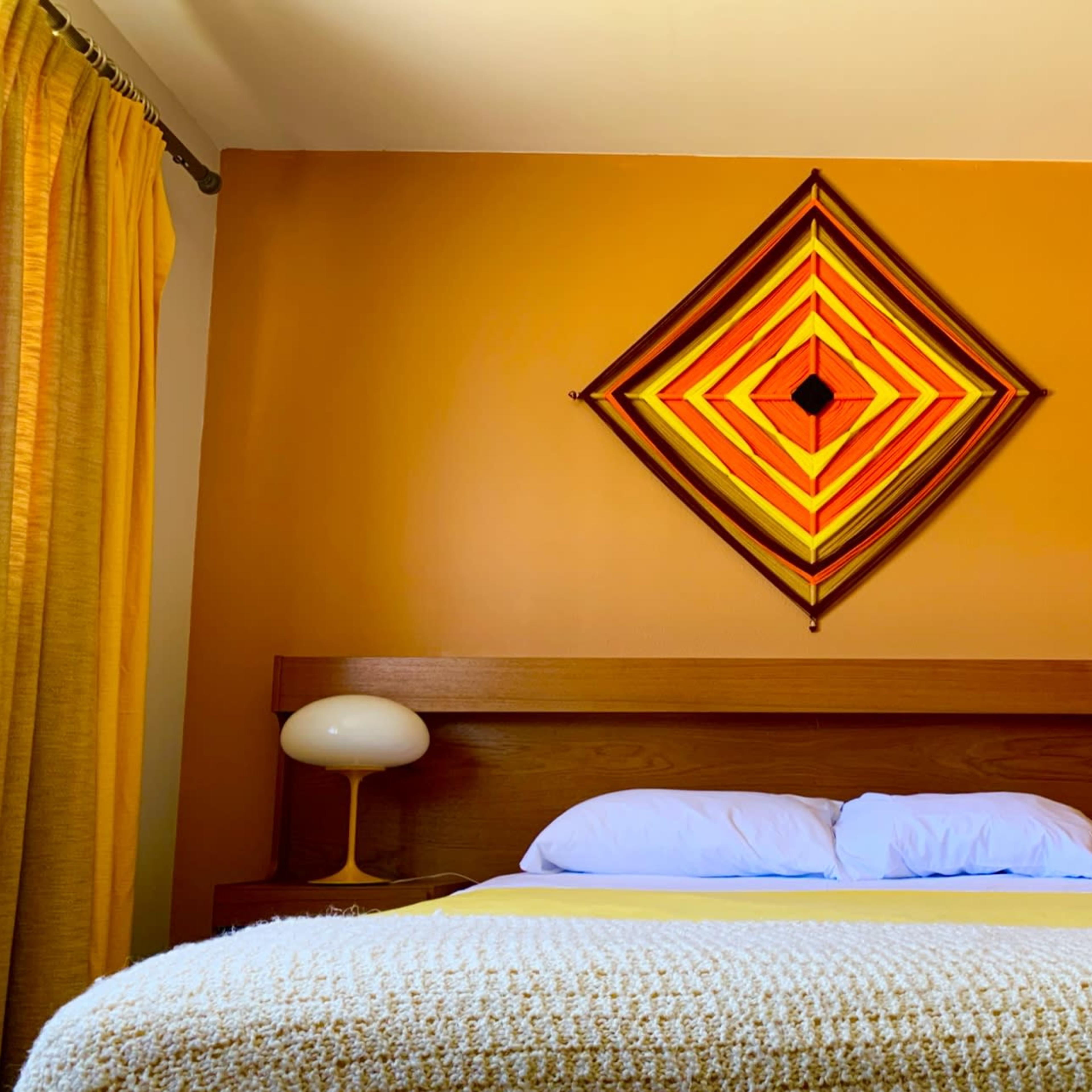 A neatly made bed with white pillows sits against a warm, orange wall, adorned with a geometric artwork, while two lamp fixtures are positioned on either side of the headboard.