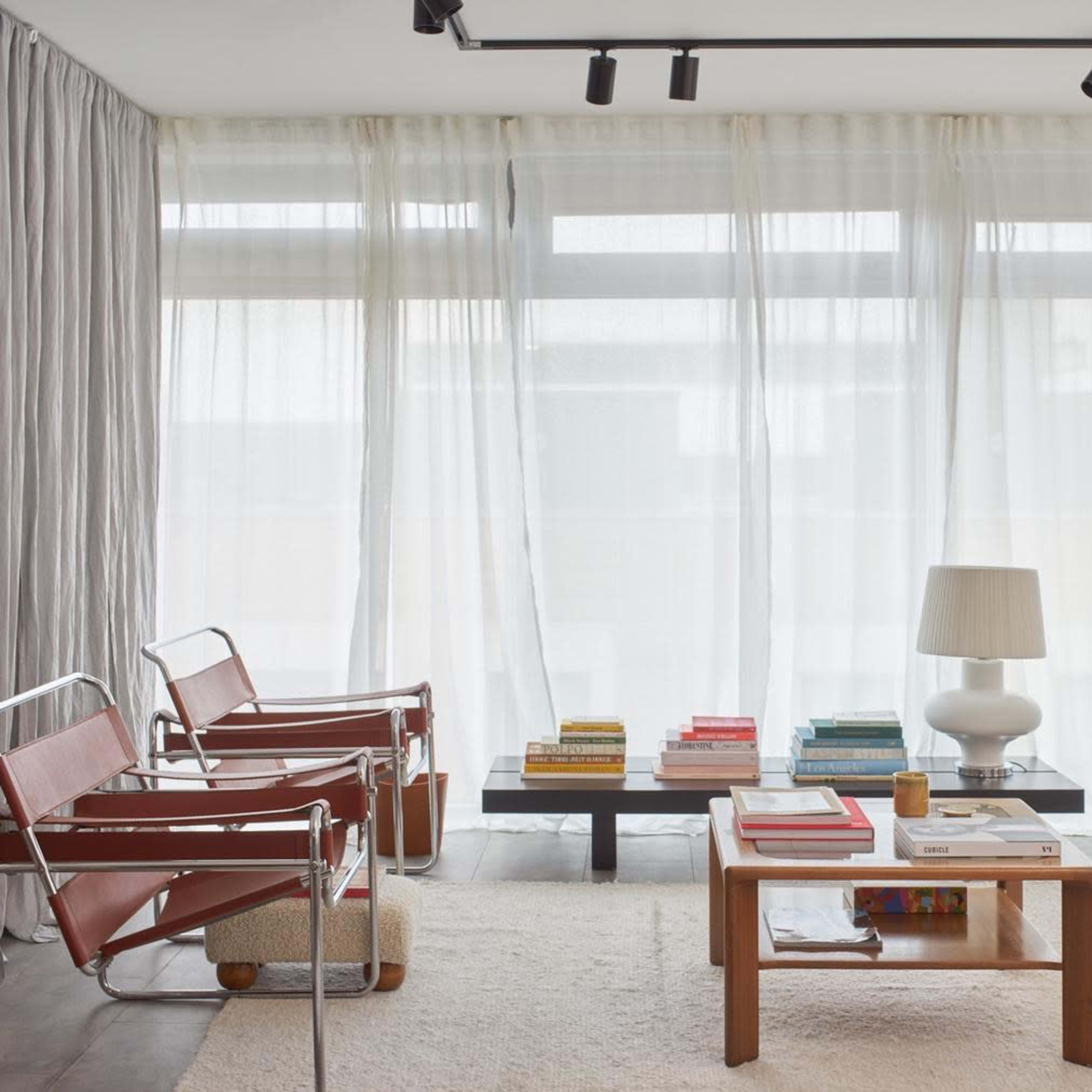 A modern living room features two metal and leather chairs, a wooden coffee table, a stack of colorful books, and a pair of upholstered armchairs, all positioned near large, sheer, white curtains.
