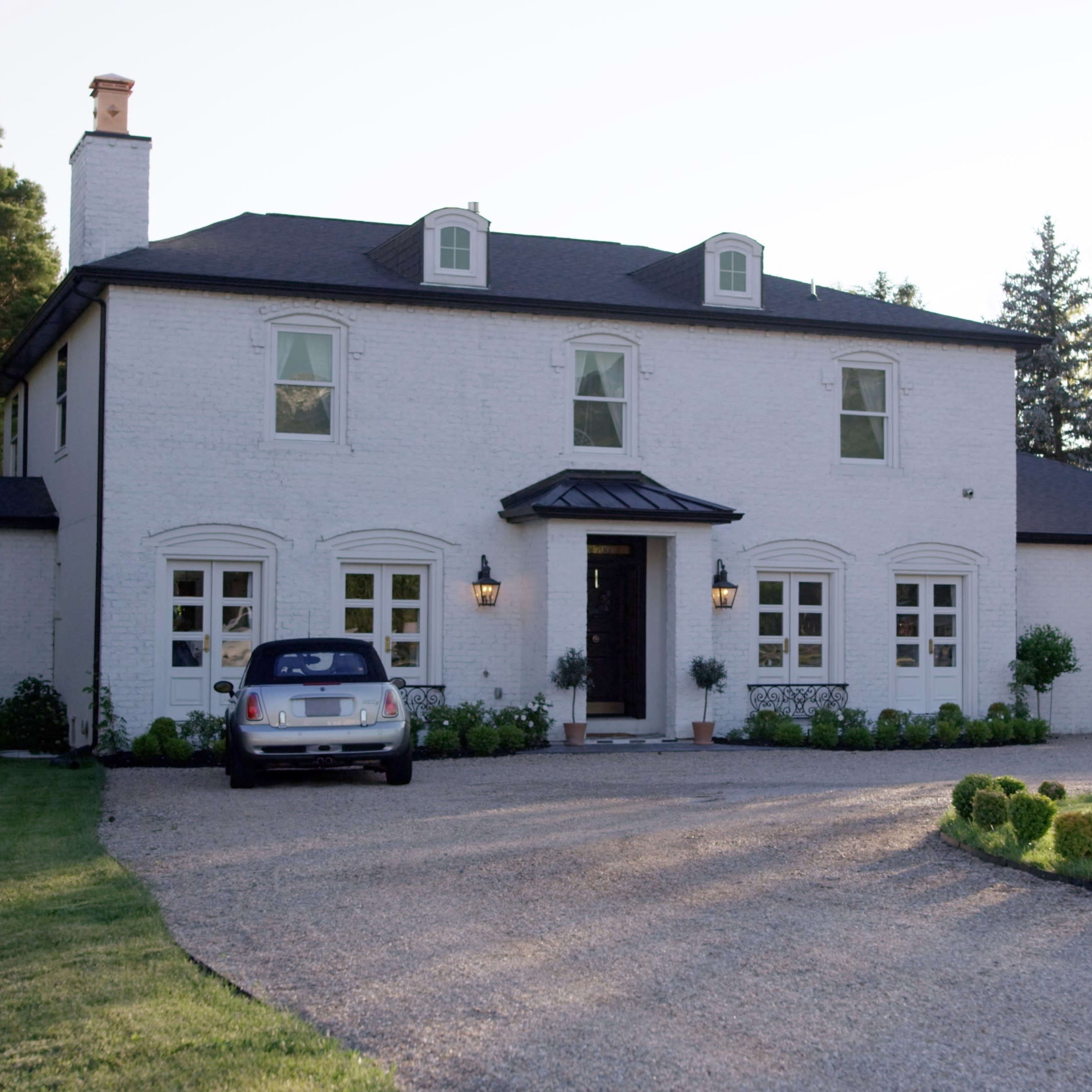 A two-story white brick house with a gravel driveway and landscaping, featuring a parked car in front.
