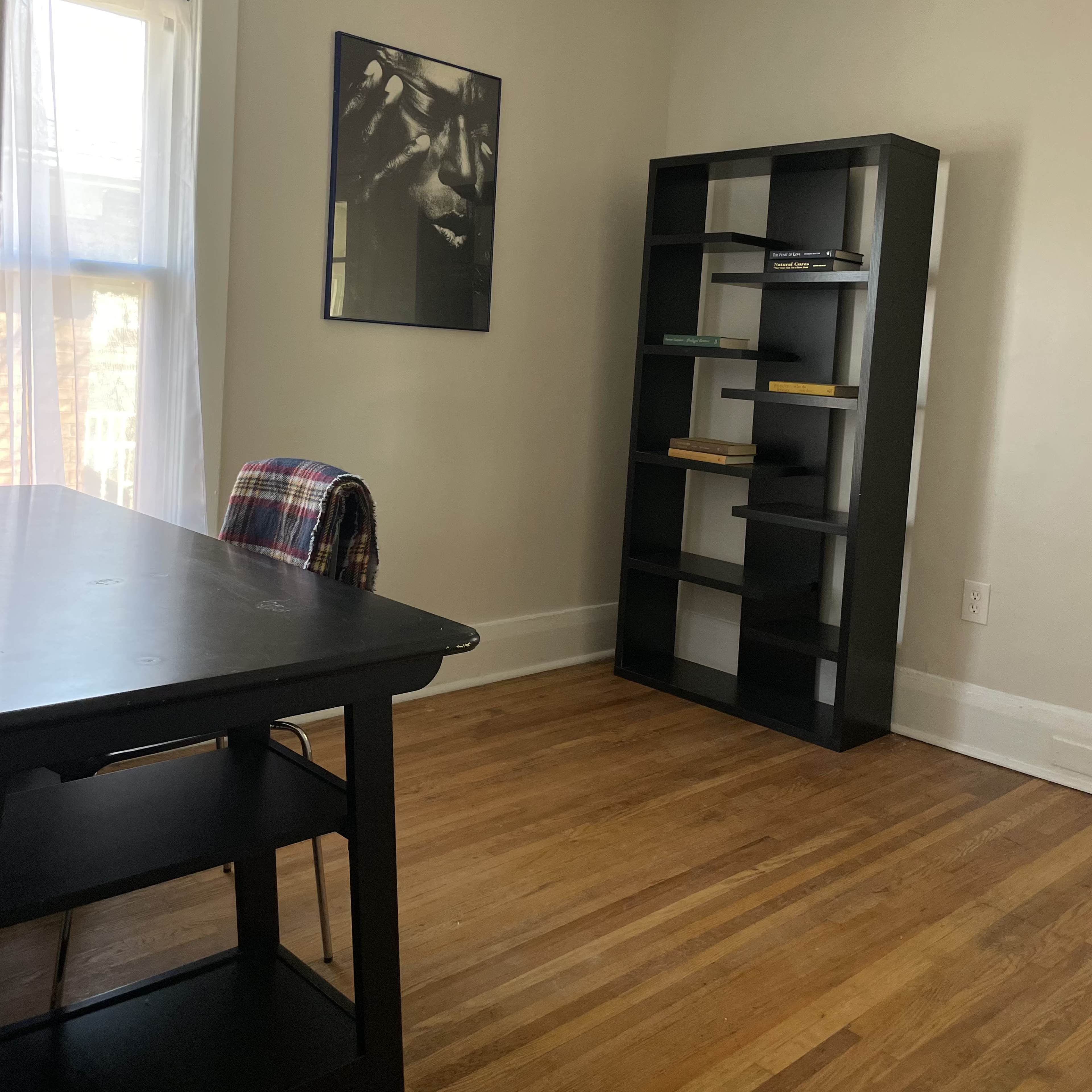A small, sparsely furnished room featuring a desk with a chair, a black bookshelf, and a wall-mounted framed picture.