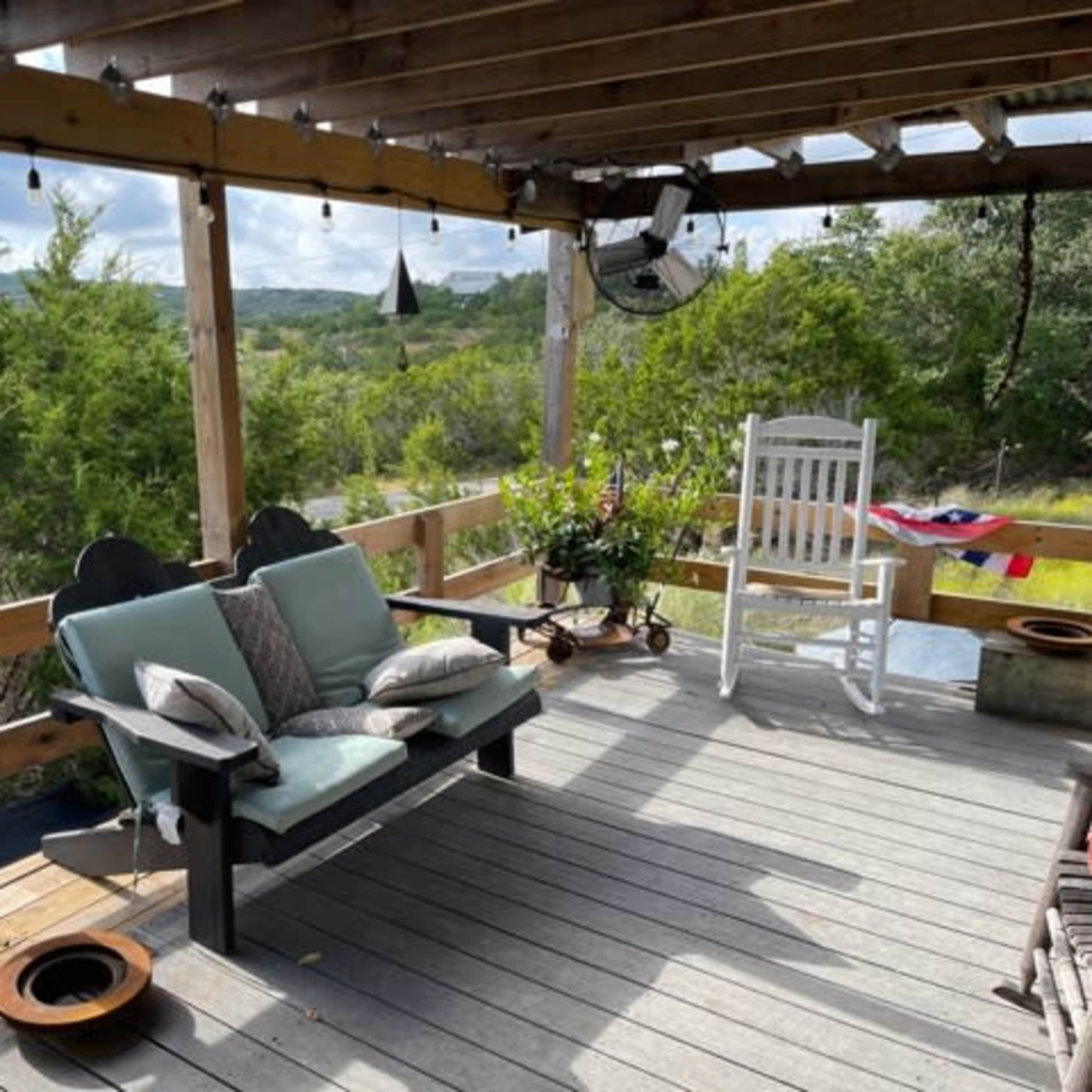 A spacious wooden deck with a swing bench, a white rocking chair, potted plants, and view of green trees in the background.