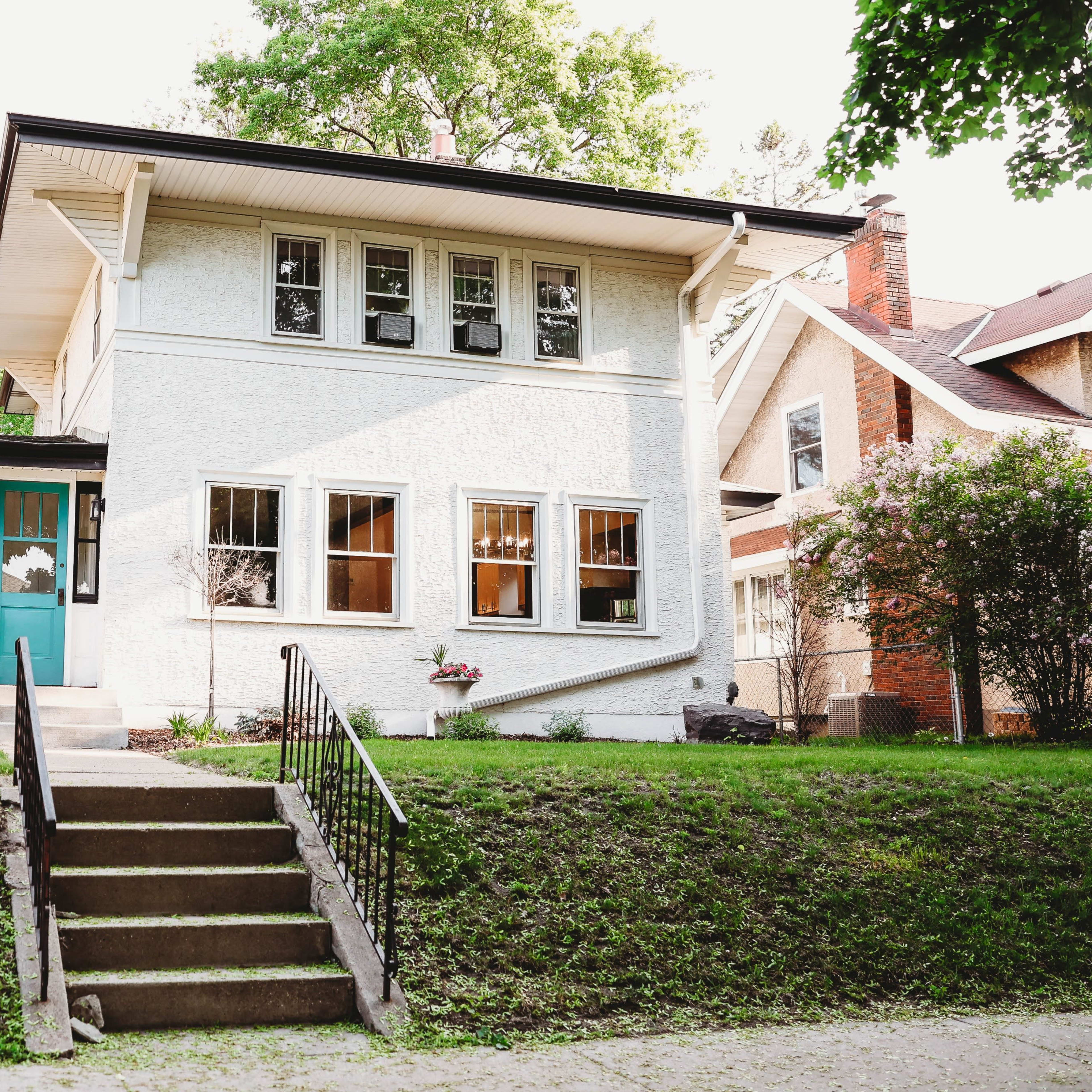 A two-story white house with a teal front door features multiple windows and is set on a landscaped yard with stone steps leading to the entrance.
