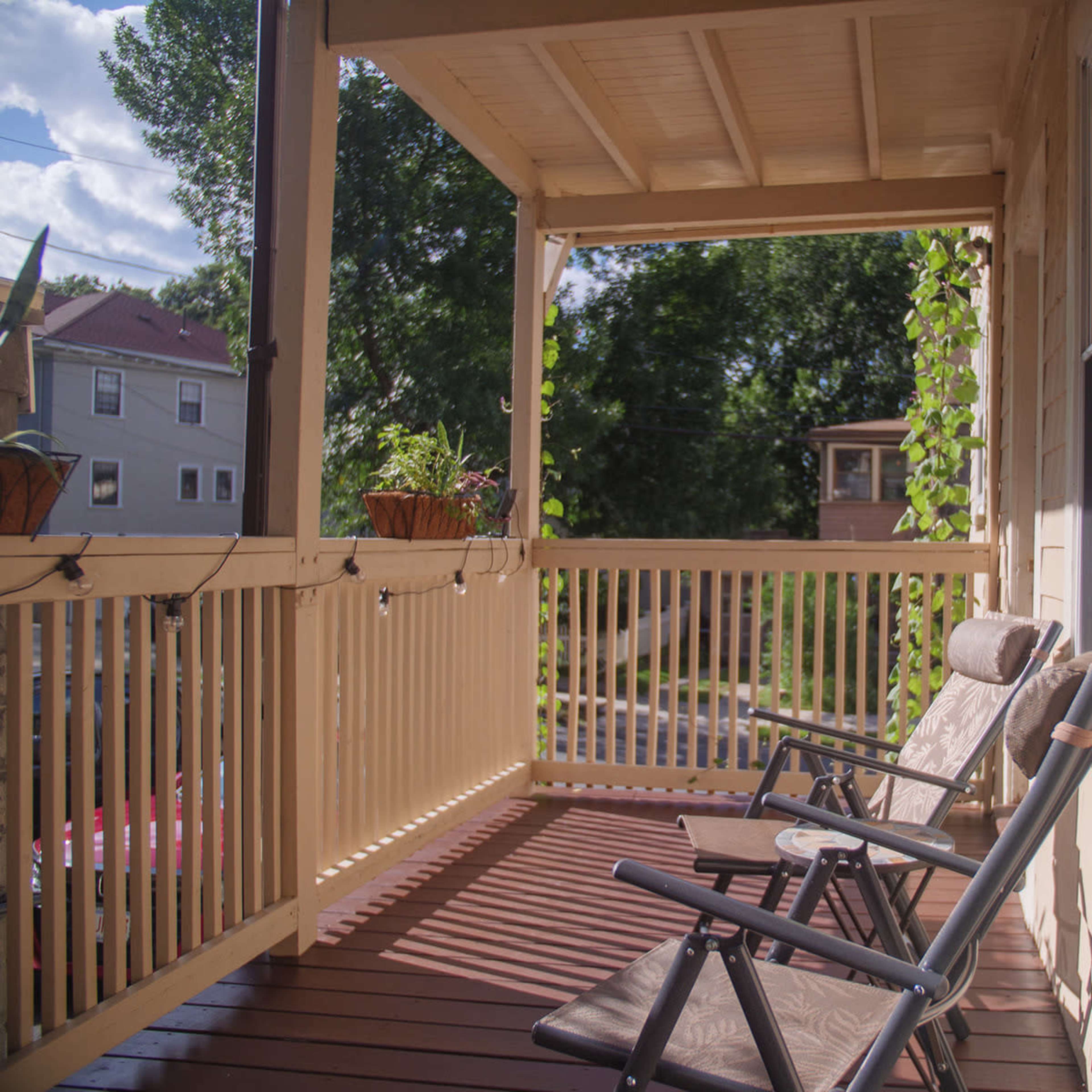 A wooden porch with two chairs and potted plants, framed by a railing and surrounded by greenery.