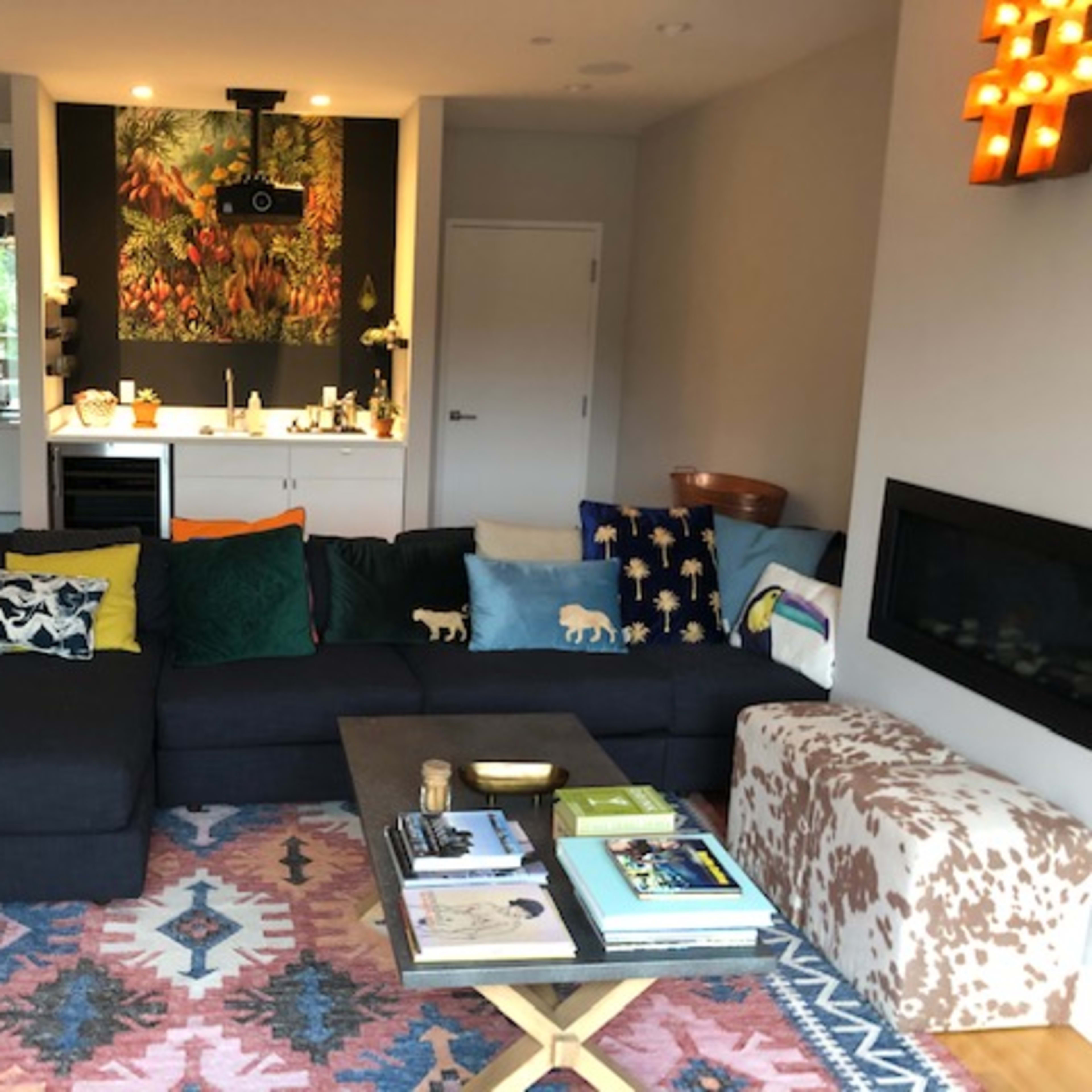 The living room features a large dark sectional sofa with vibrant throw pillows, a coffee table stacked with books, and a colorful area rug underfoot, all set against a backdrop of modern decor.