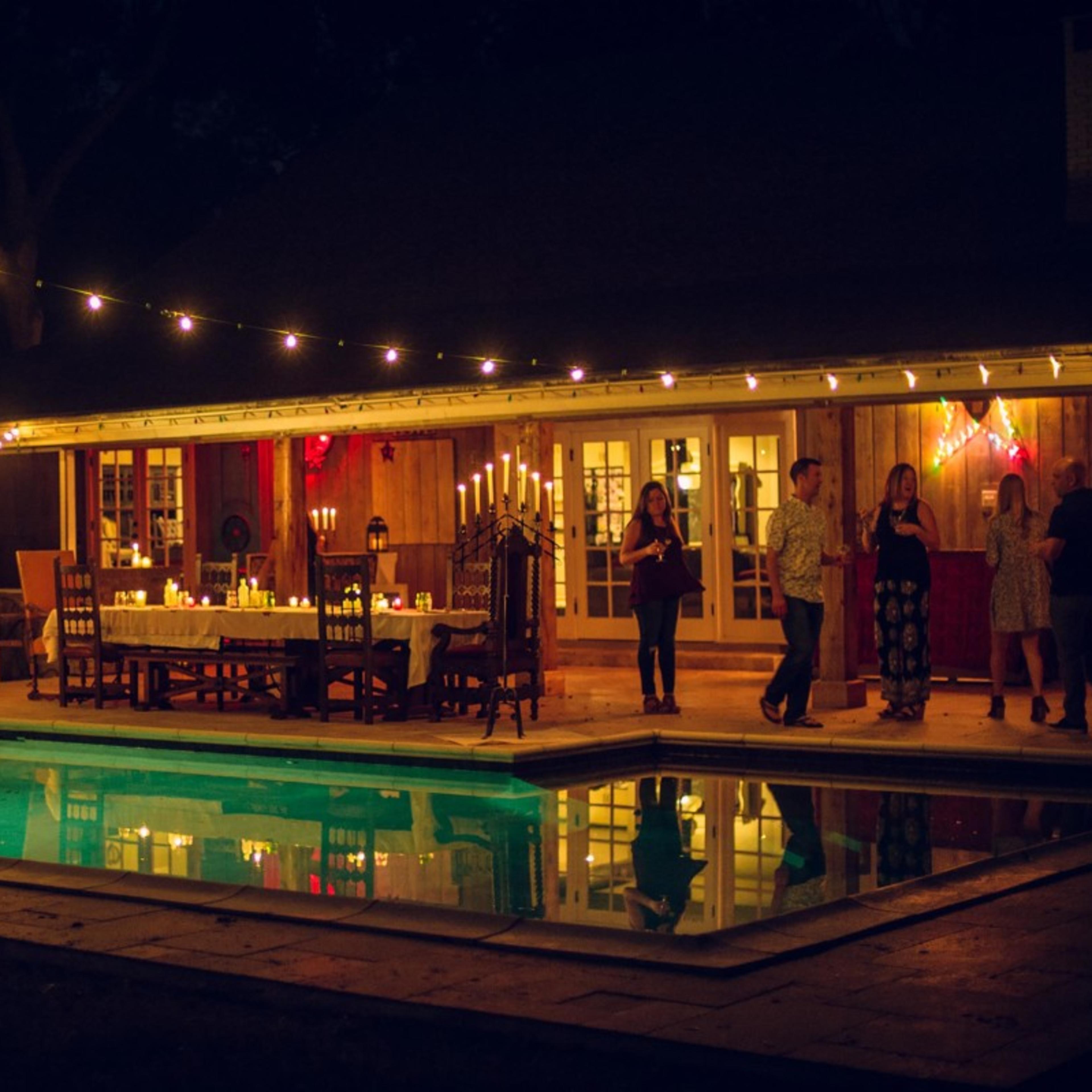A beautifully set outdoor dining area is illuminated by string lights beside a swimming pool at night.