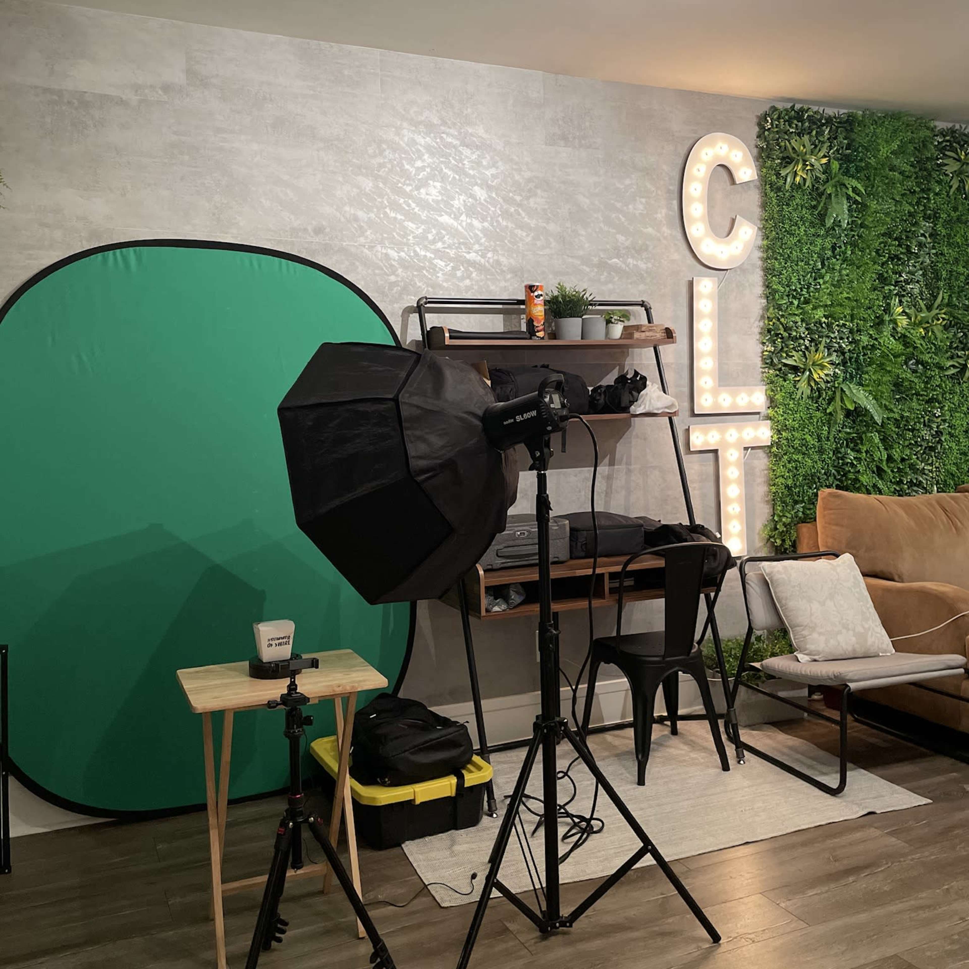 A photography setup with a green backdrop, a softbox light, and a shelf featuring decorative items against a wall with greenery and illuminated letters spelling "CLT."