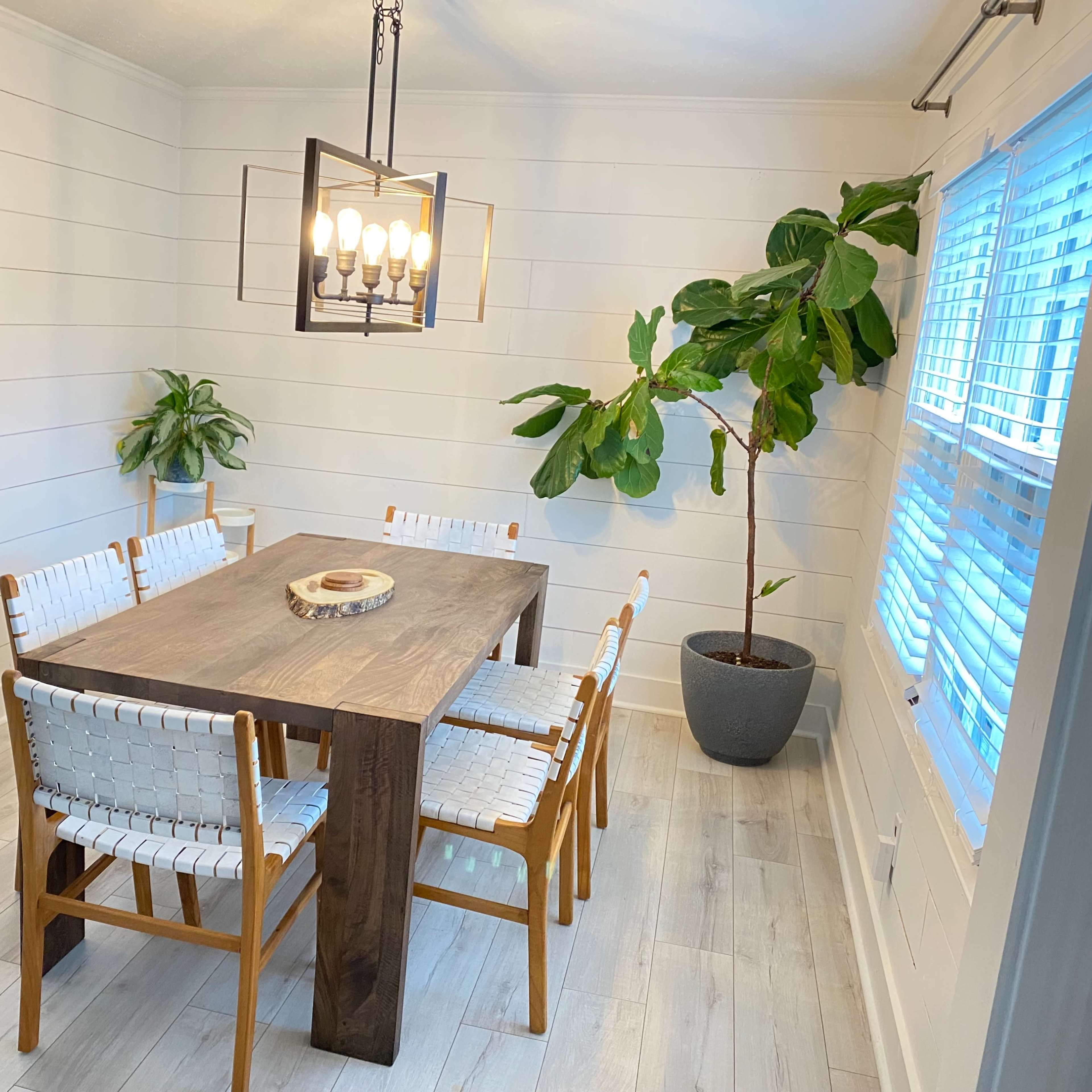 A dining room features a wooden table surrounded by six white woven chairs, with two potted plants near a window.