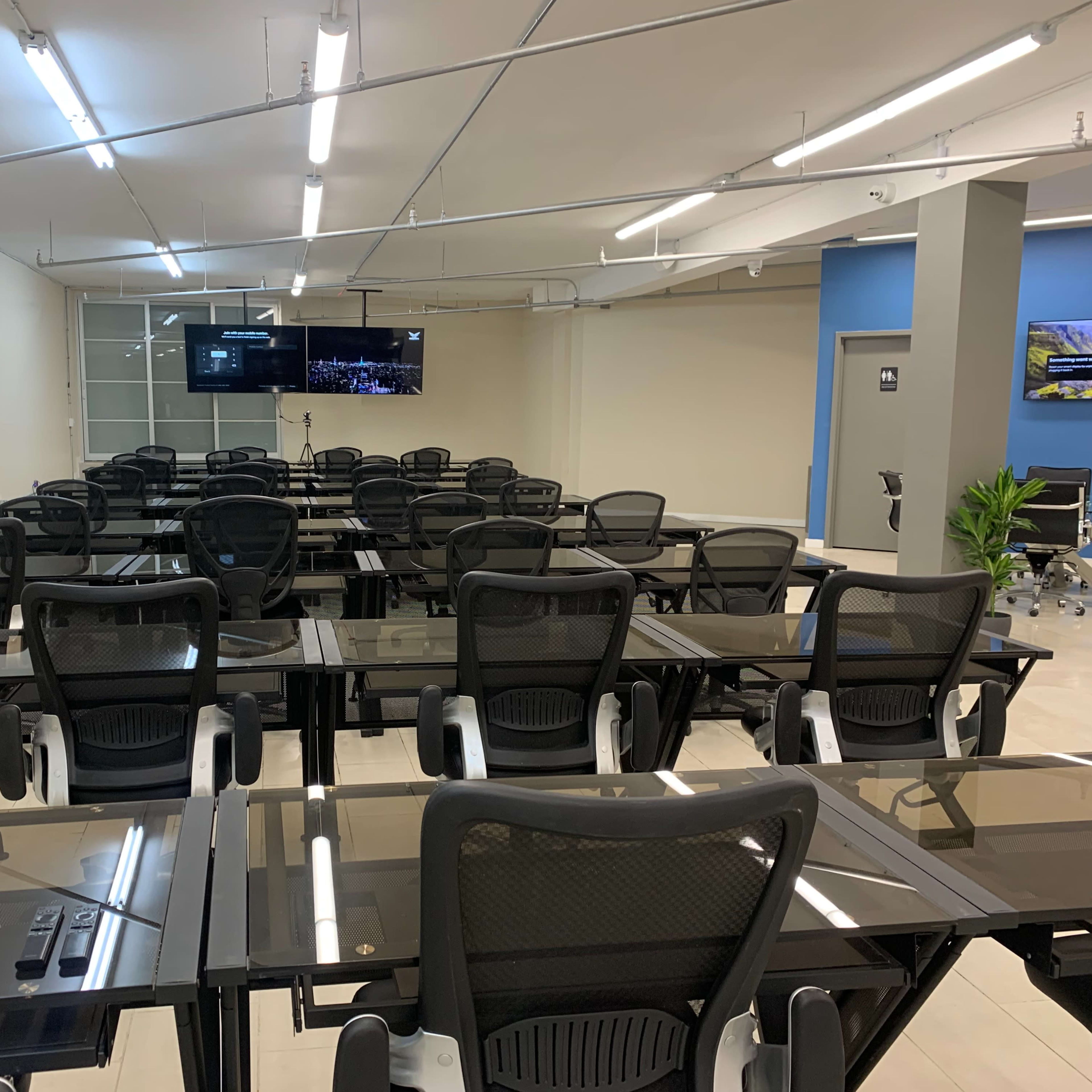A classroom is set up with rows of glass-top desks and black mesh chairs, facing two screens on the front wall.
