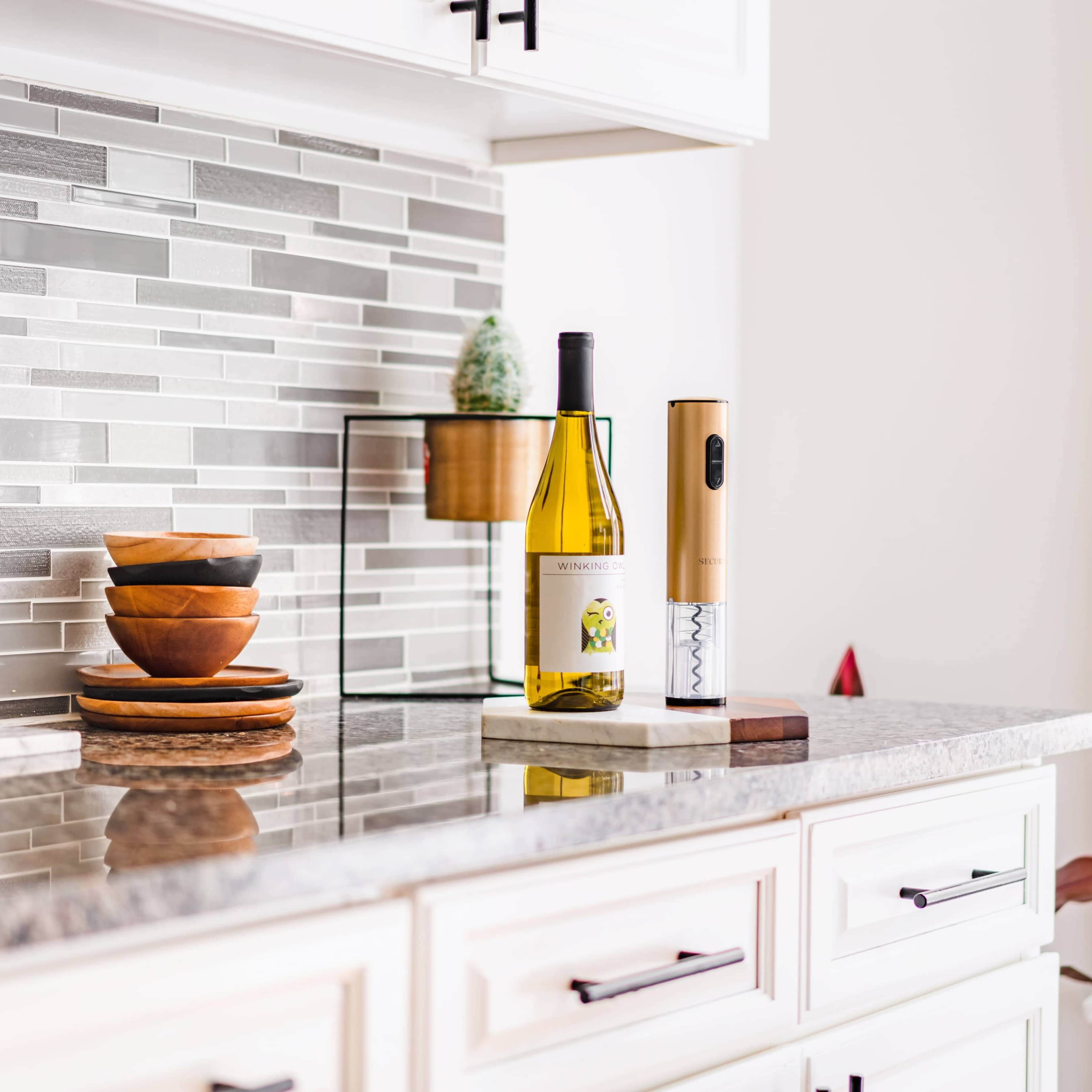 A countertop features a wine bottle, an electric corkscrew, and bowls stacked beside a decorative plant on a tiled backsplash.