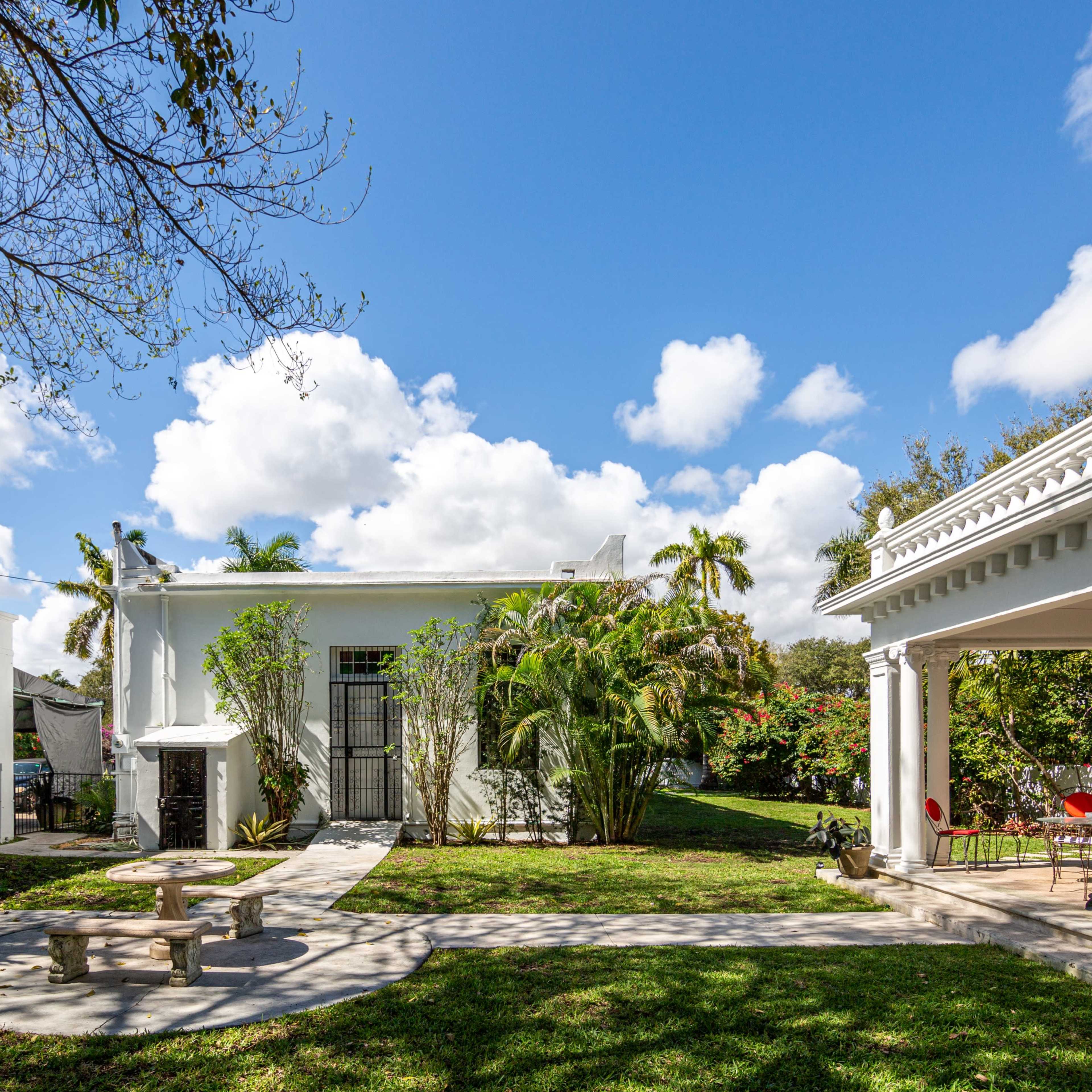 A landscaped outdoor area featuring a white building, a pavilion with columns, and a circular stone bench surrounded by greenery under a partly cloudy sky.