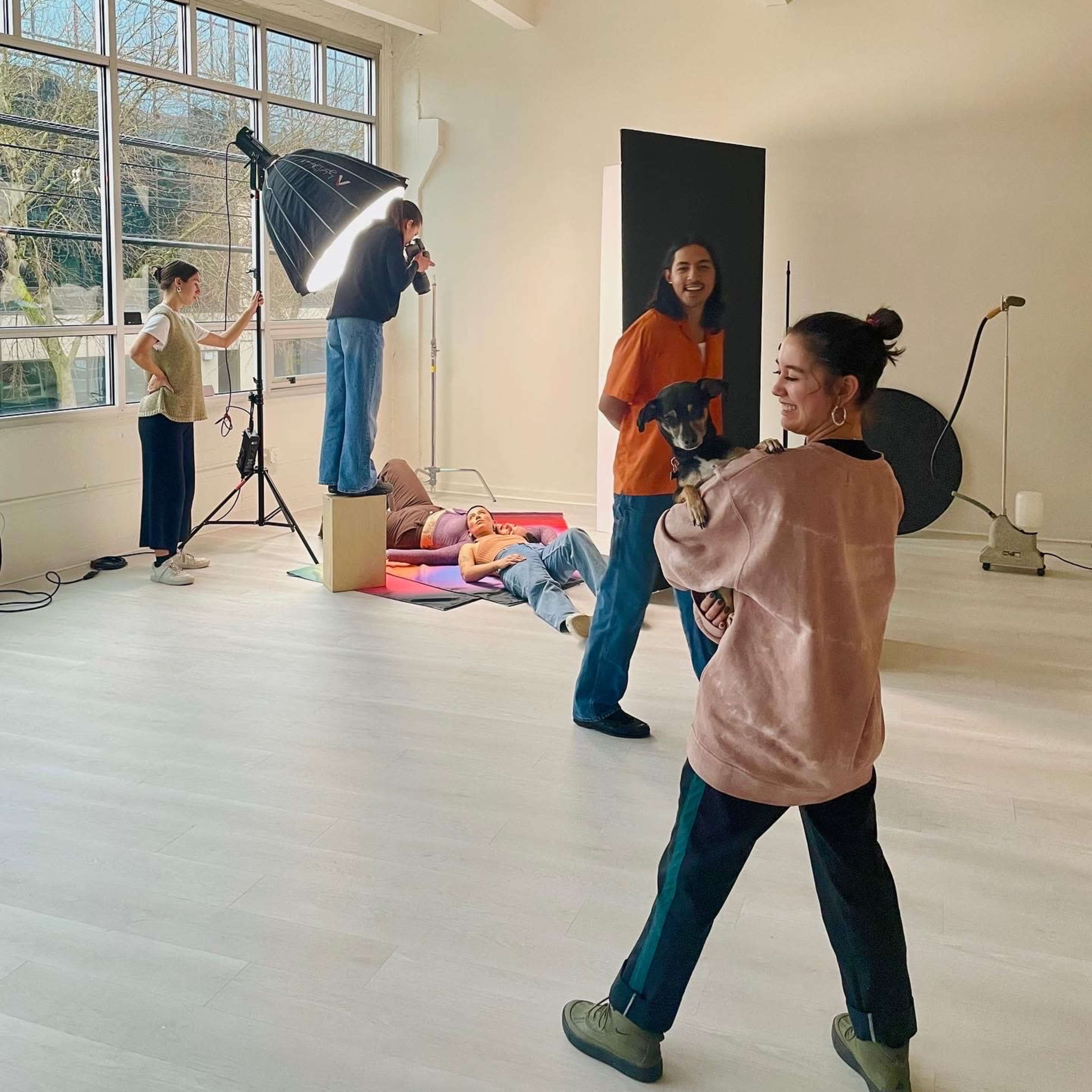 A group of people is engaged in a photoshoot in a well-lit studio, with one person posing on a box while others adjust lighting and equipment.
