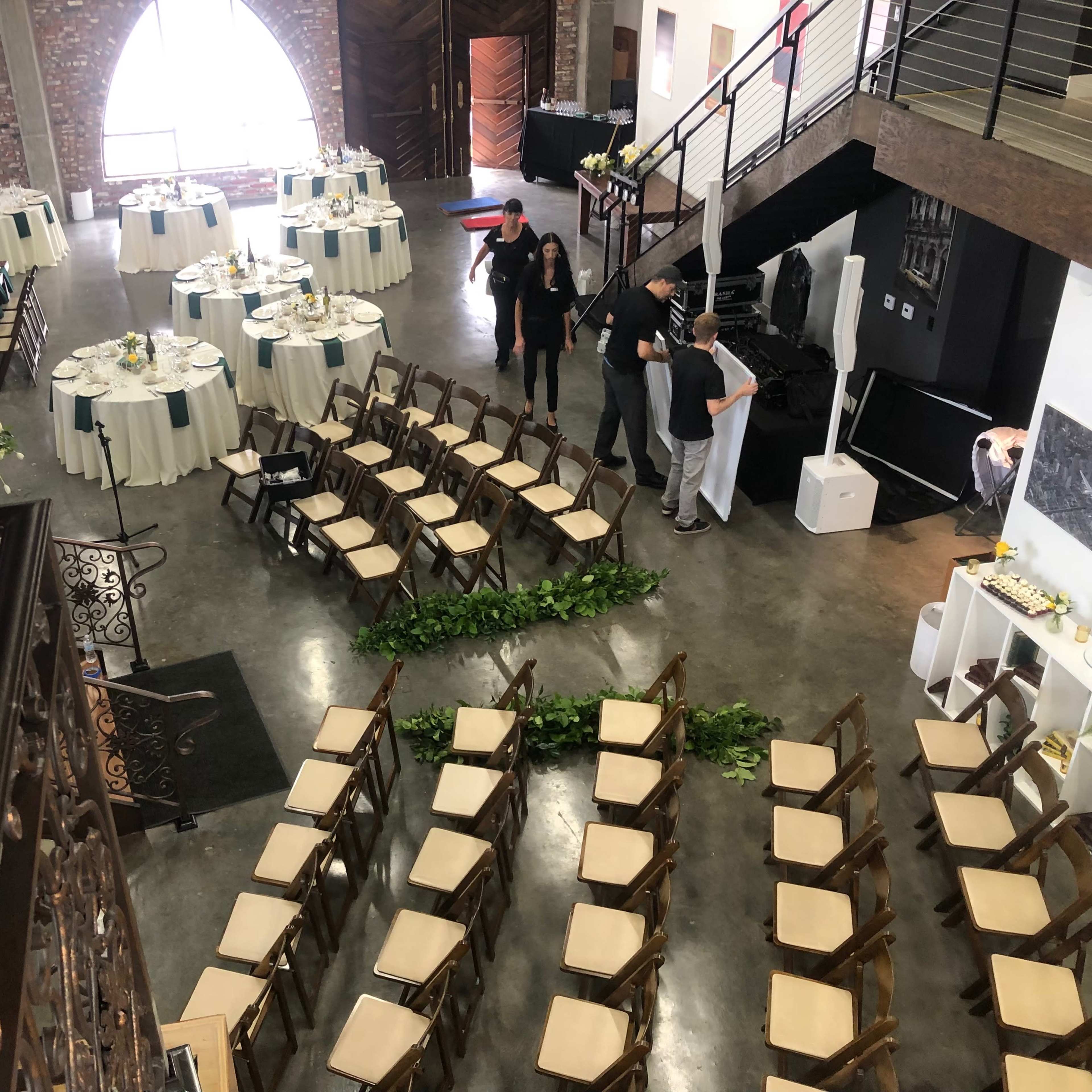 A multi-level event space with dining tables arranged for a reception and chairs set up for a ceremony beneath large arched windows.