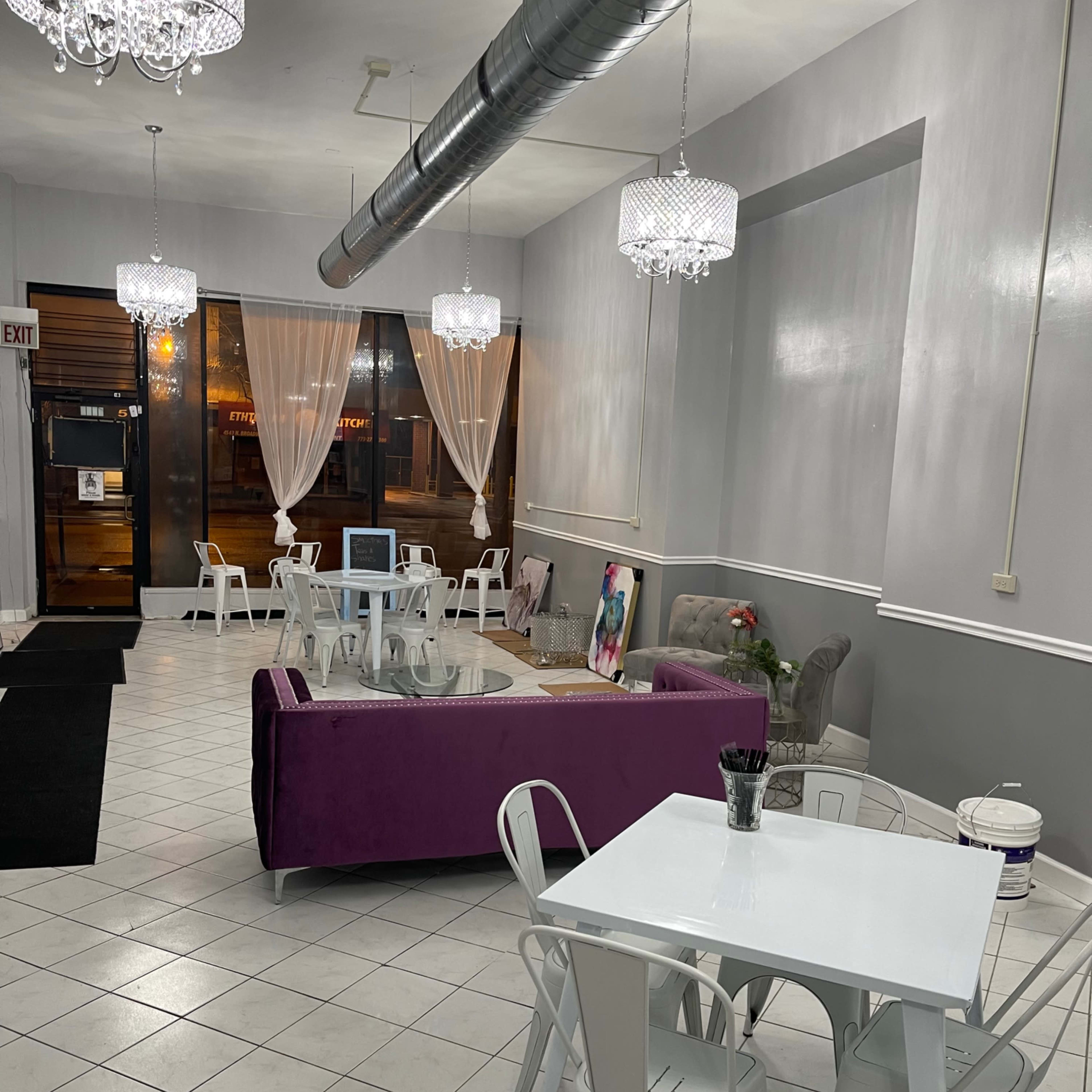 A spacious cafe with white tile flooring, a purple couch, white tables, and decorative chandeliers.
