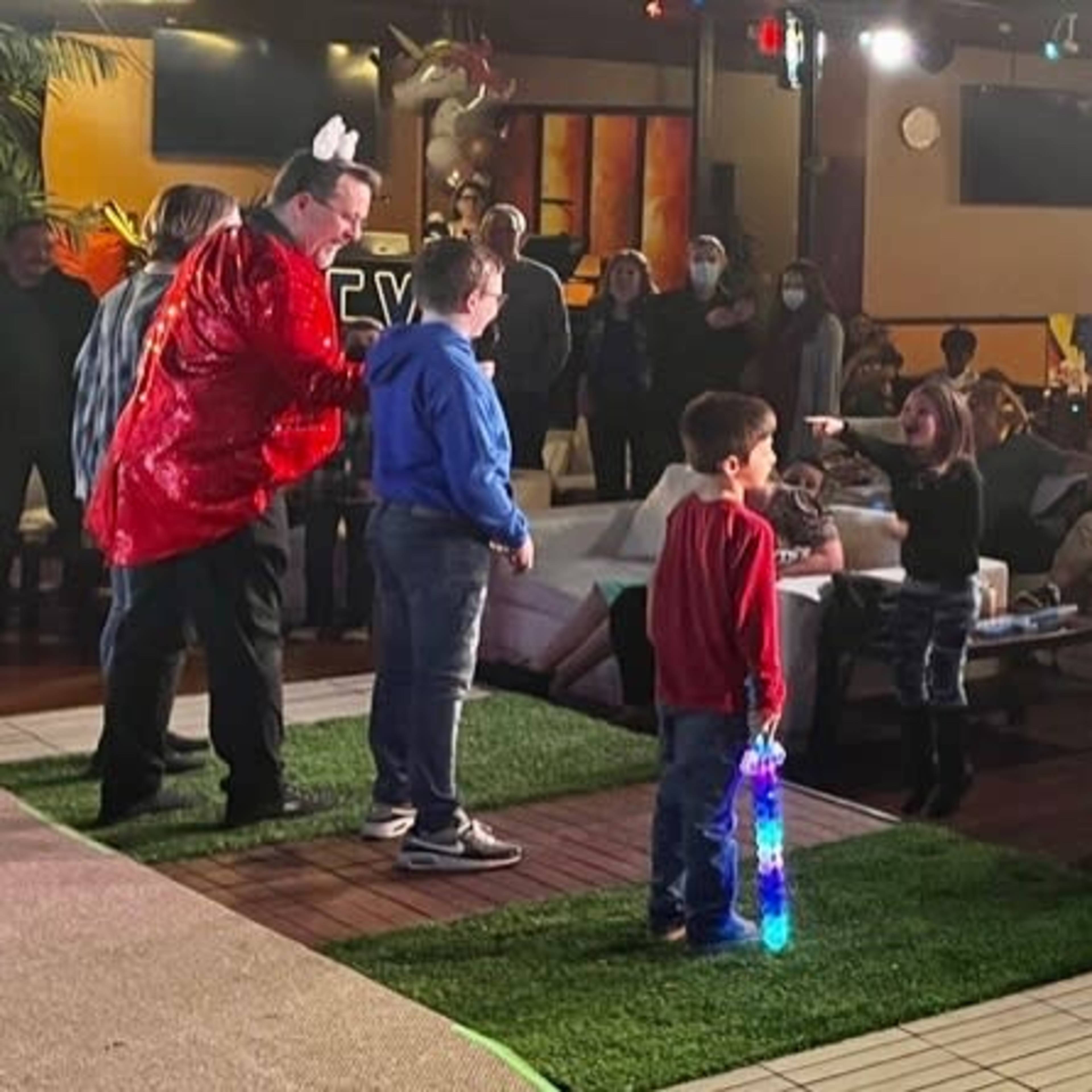 A lively event is taking place in a spacious venue filled with guests of various ages, some seated at tables while others participate in activities on a stage area with colorful lighting.