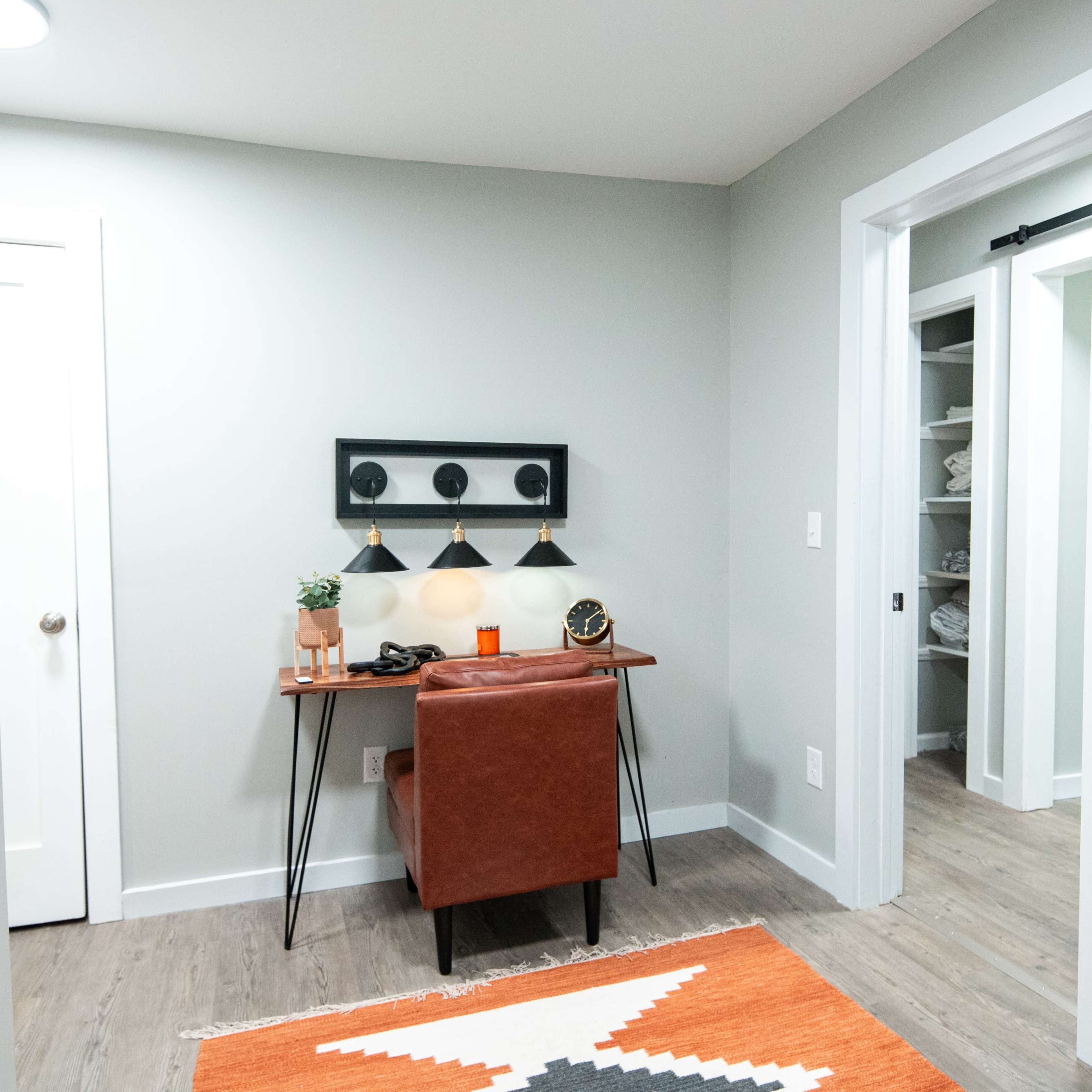A small entryway with a brown leather chair and a table against a wall, featuring three light fixtures above it, a plant, and a rug on the floor.