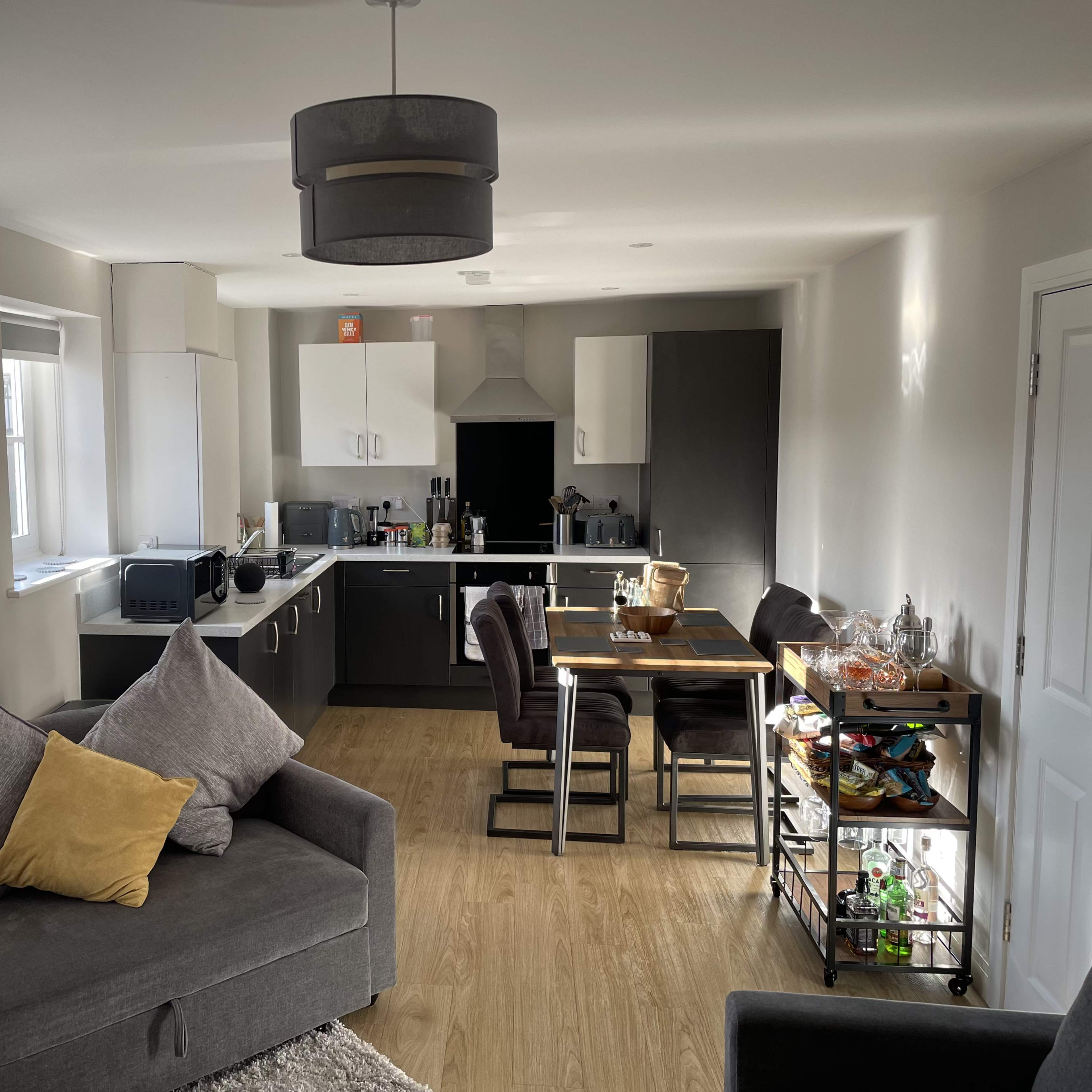 A modern open-plan living space featuring a gray sectional sofa, a kitchen with dark cabinetry, and a dining area with a wooden table and chairs.