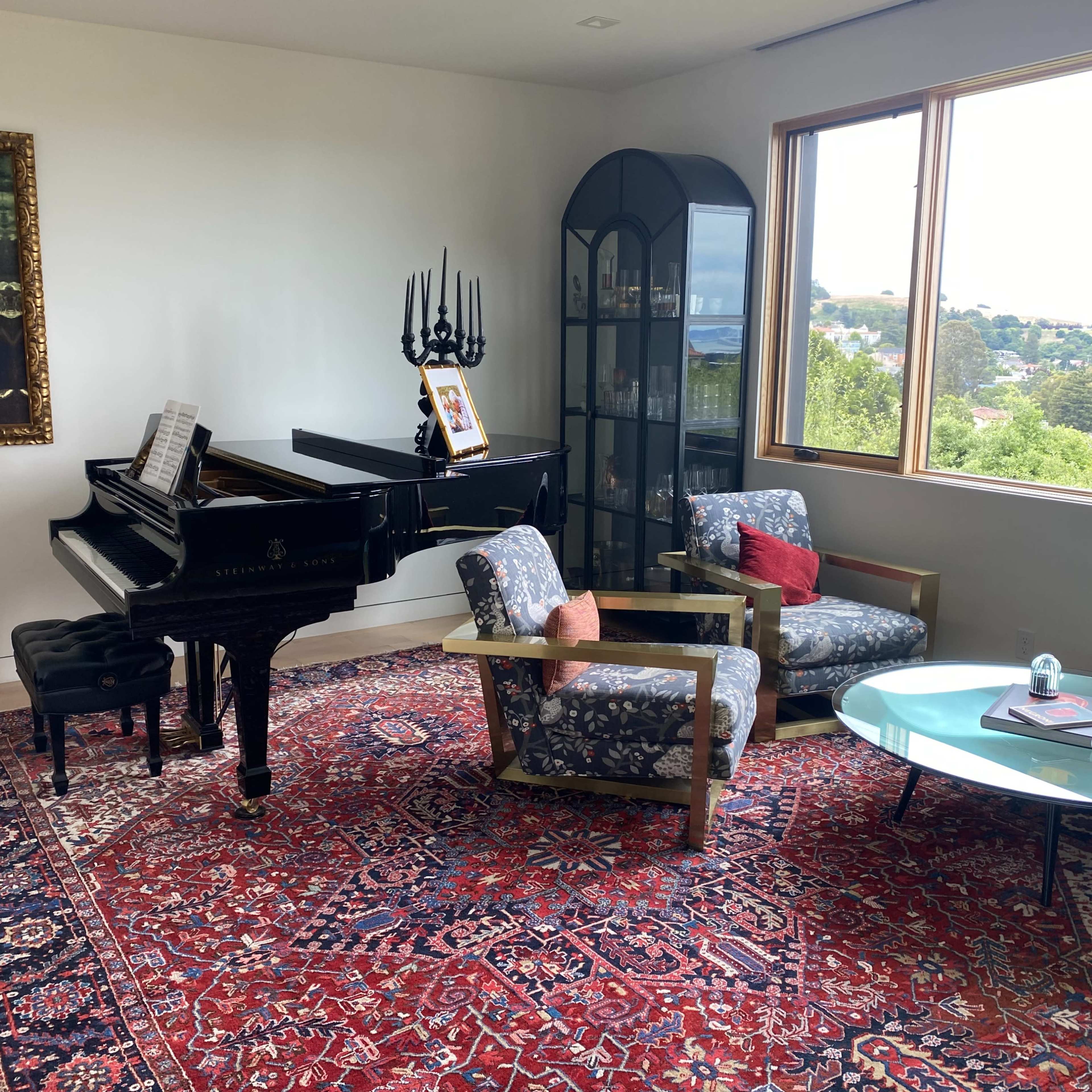 A grand piano sits next to two patterned chairs on a large rug in a room with a large window overlooking a hillside view.