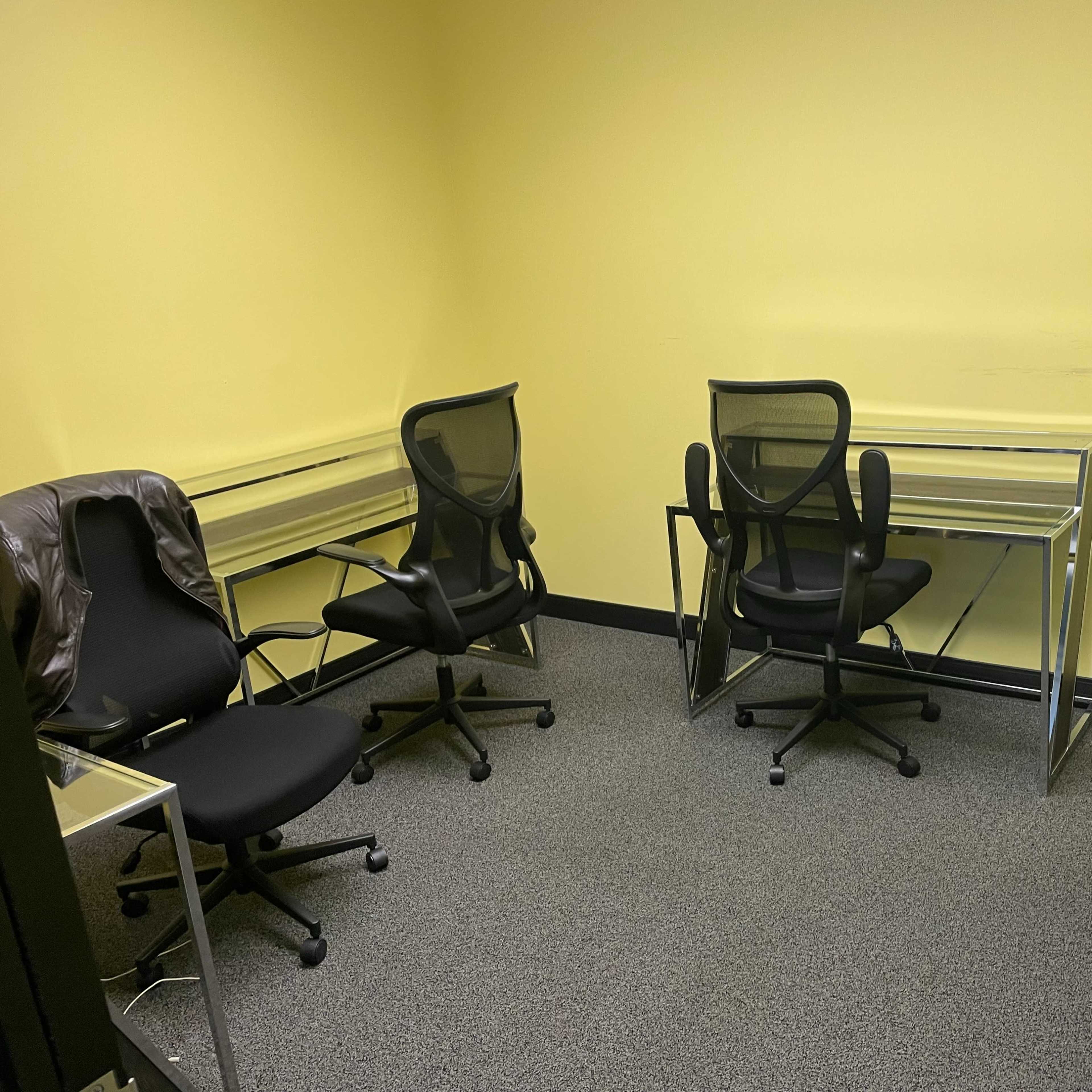 An empty room with yellow walls, containing several black office chairs and glass-top desks arranged in a circular layout.