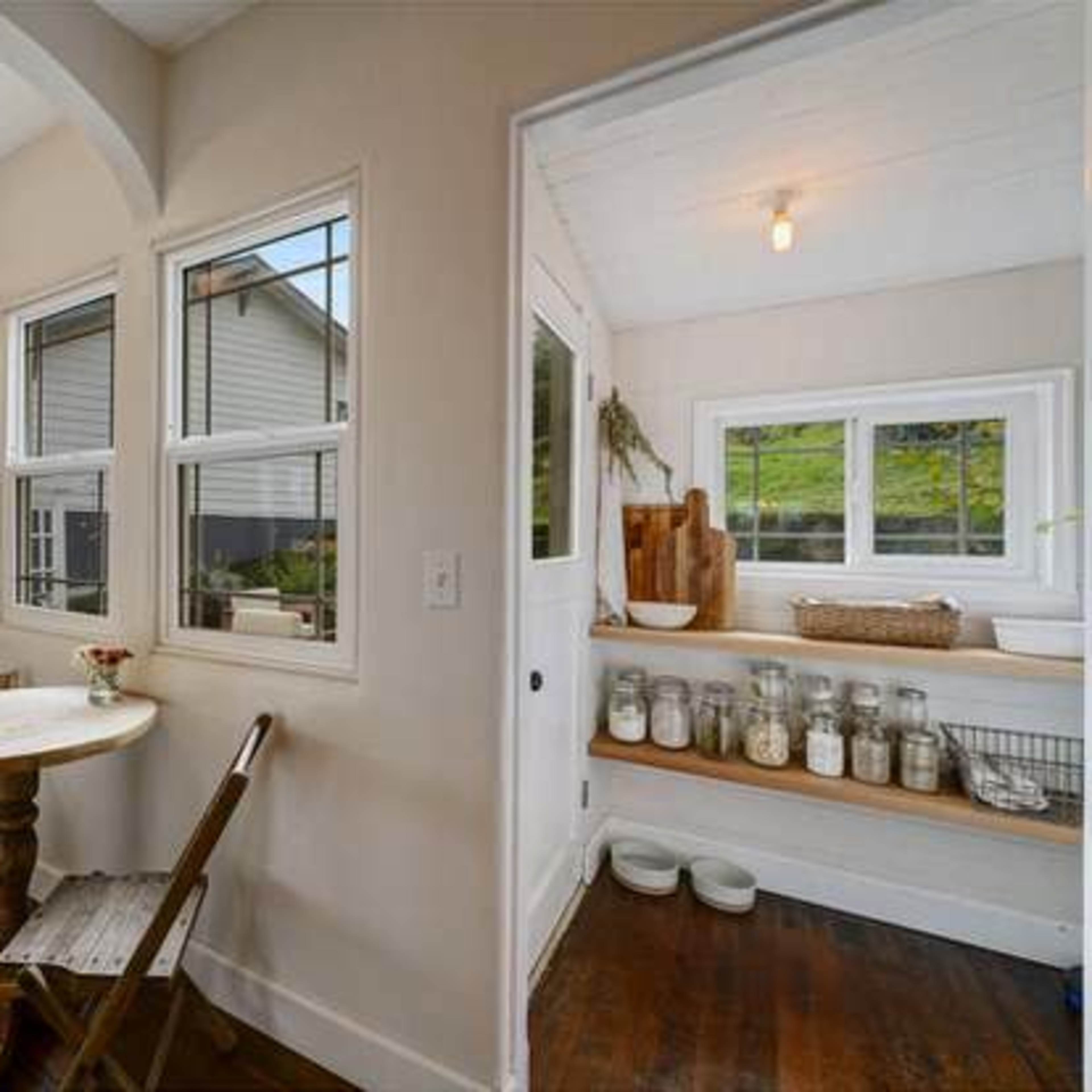 A small kitchen space features a round table with a single chair, a chalkboard on the wall, and a windowed nook displaying jars and a wooden cutting board.