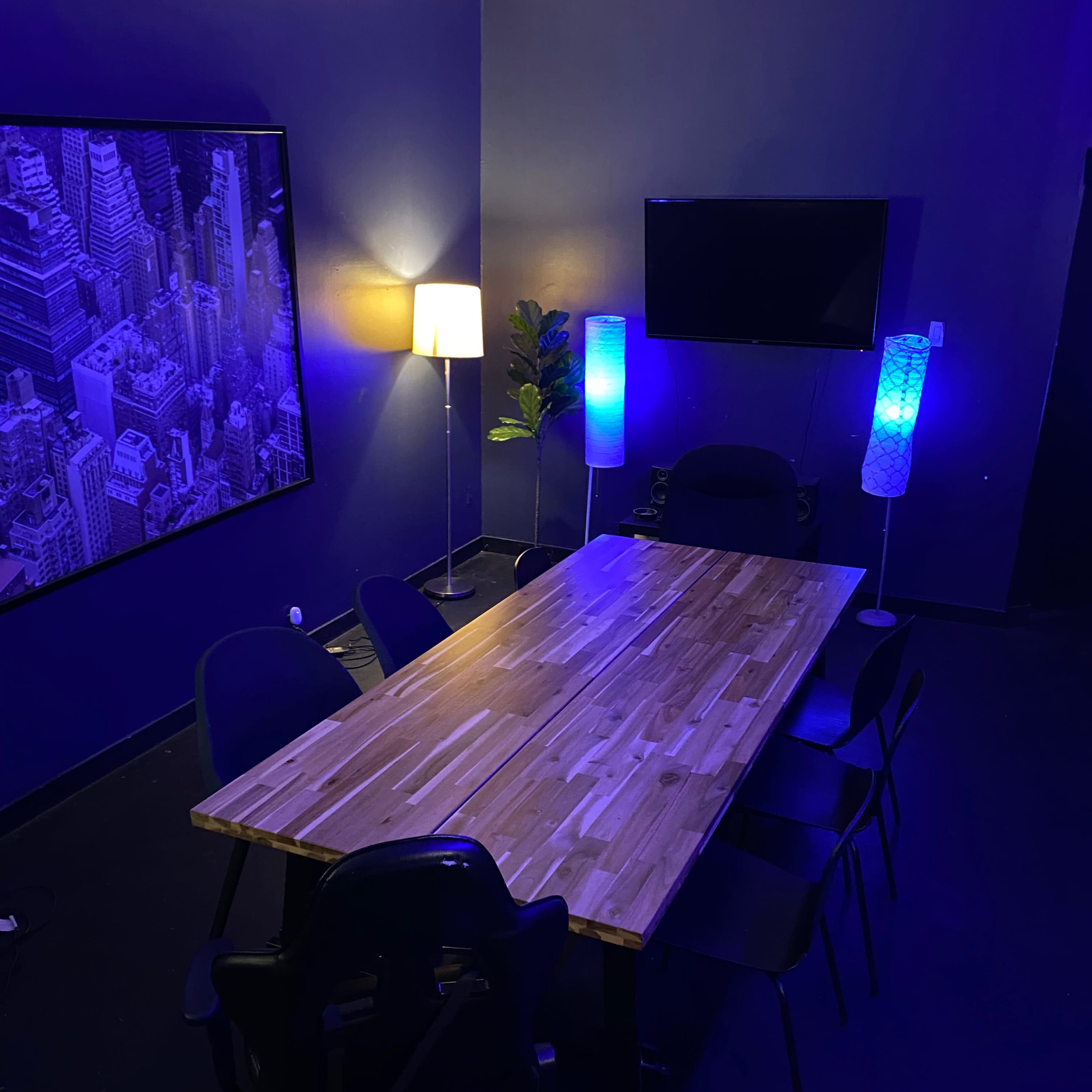 A large wooden conference table surrounded by black chairs is set in a dimly lit room with blue-and-green lamps and a wall-mounted television.