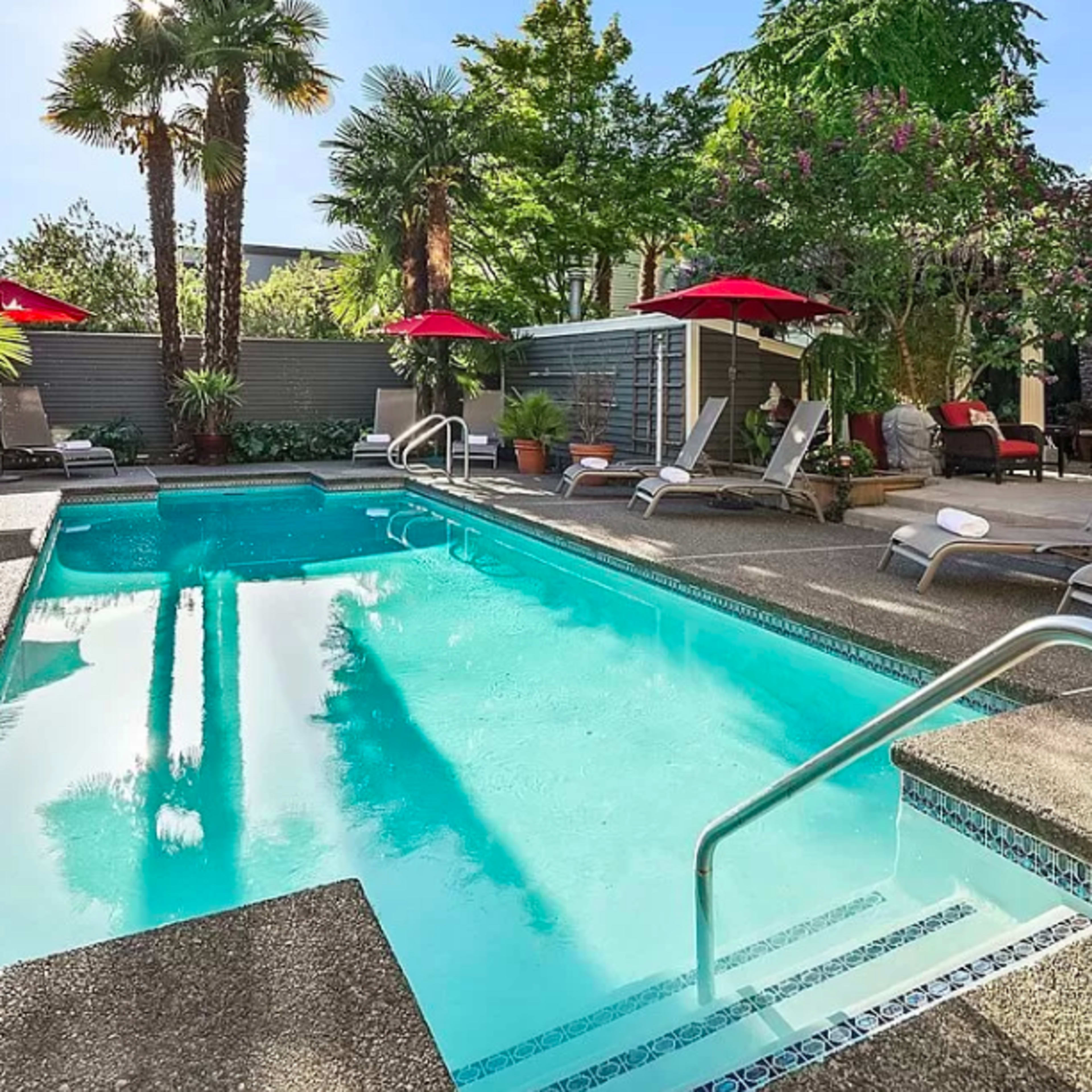 A swimming pool surrounded by palm trees and lounge chairs, with red umbrellas providing shade.