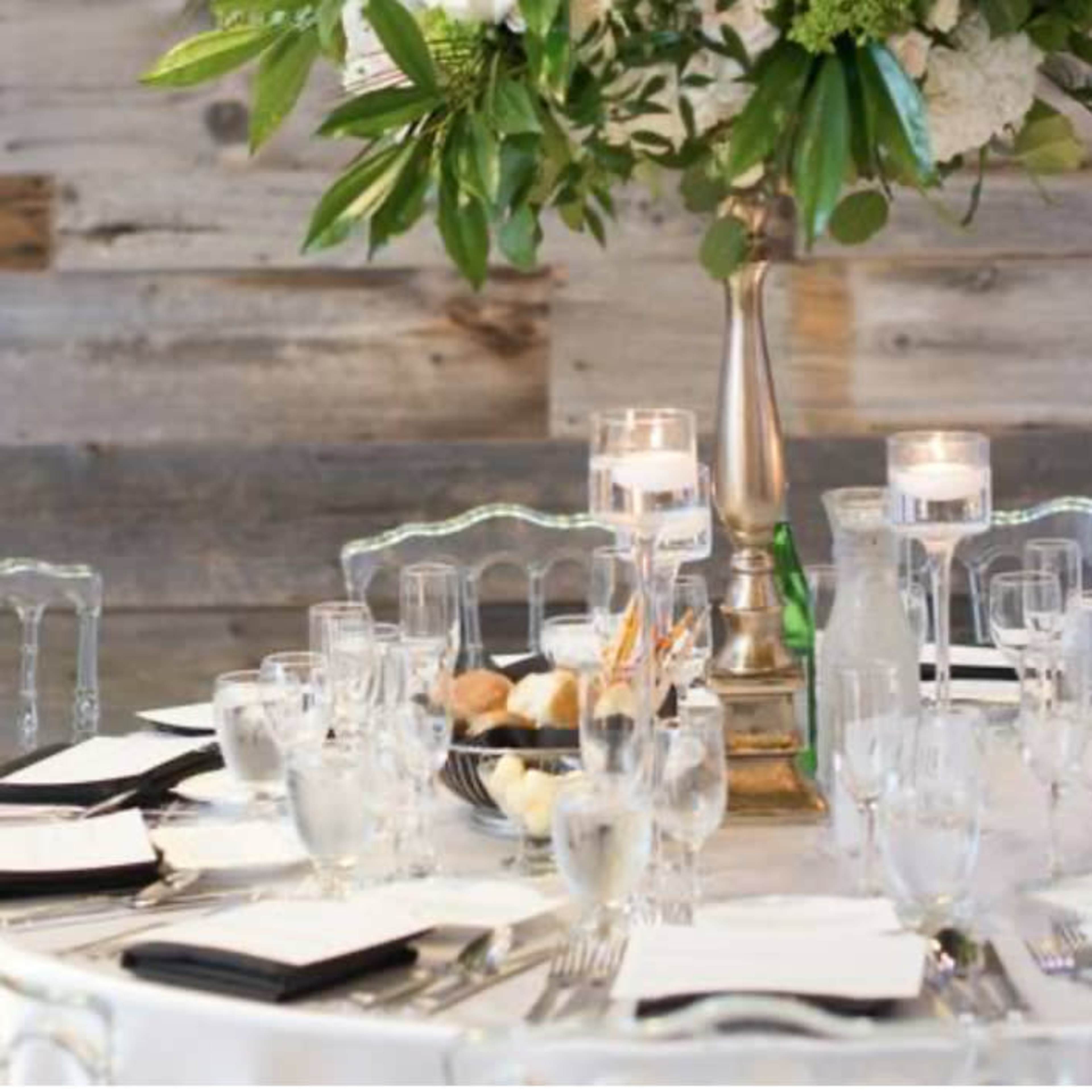 A round dining table is elegantly set with crystal glassware, rolled napkins, and a tall floral centerpiece, surrounded by clear chairs.