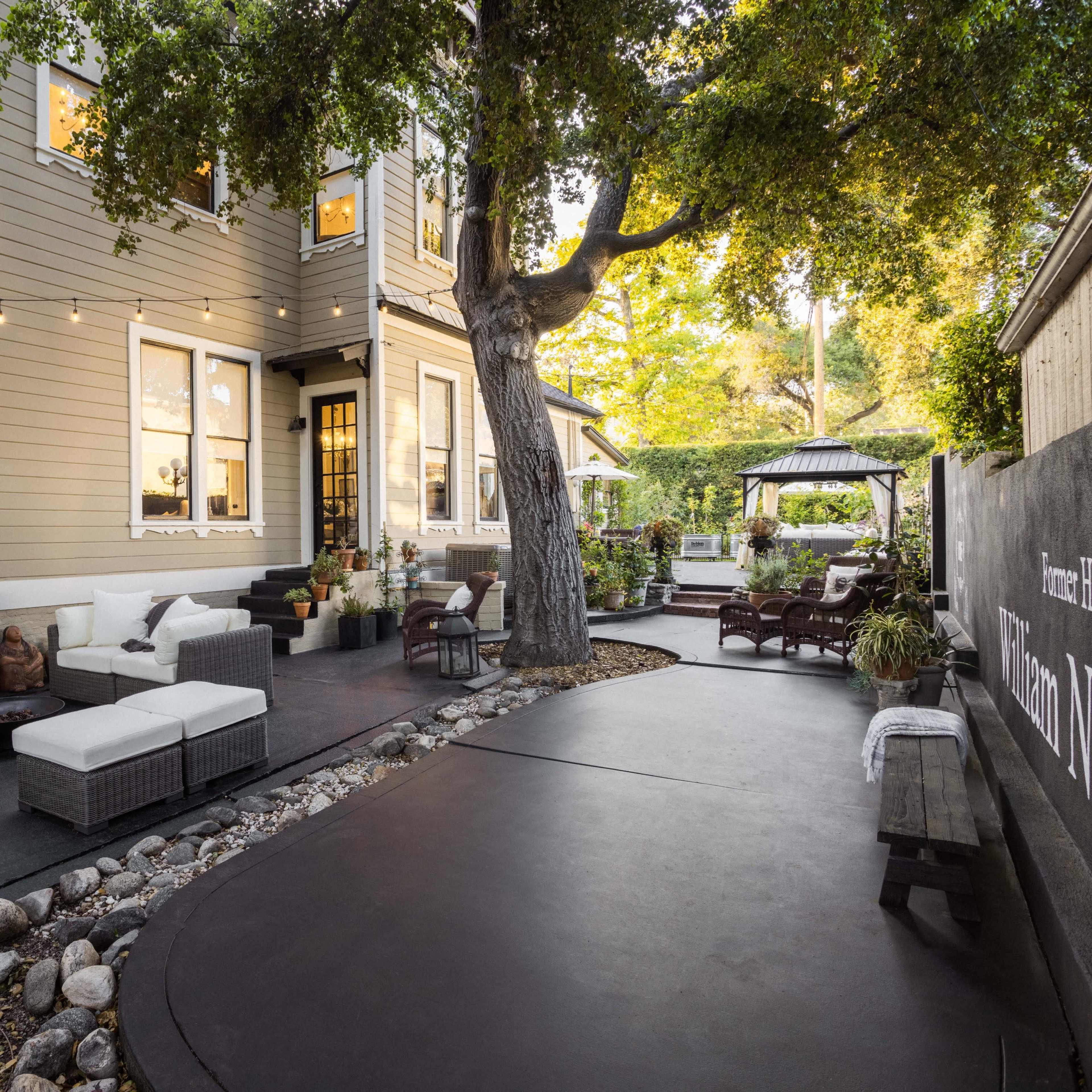 A landscaped backyard with seating areas, stone pathways, and a large tree beside a house.