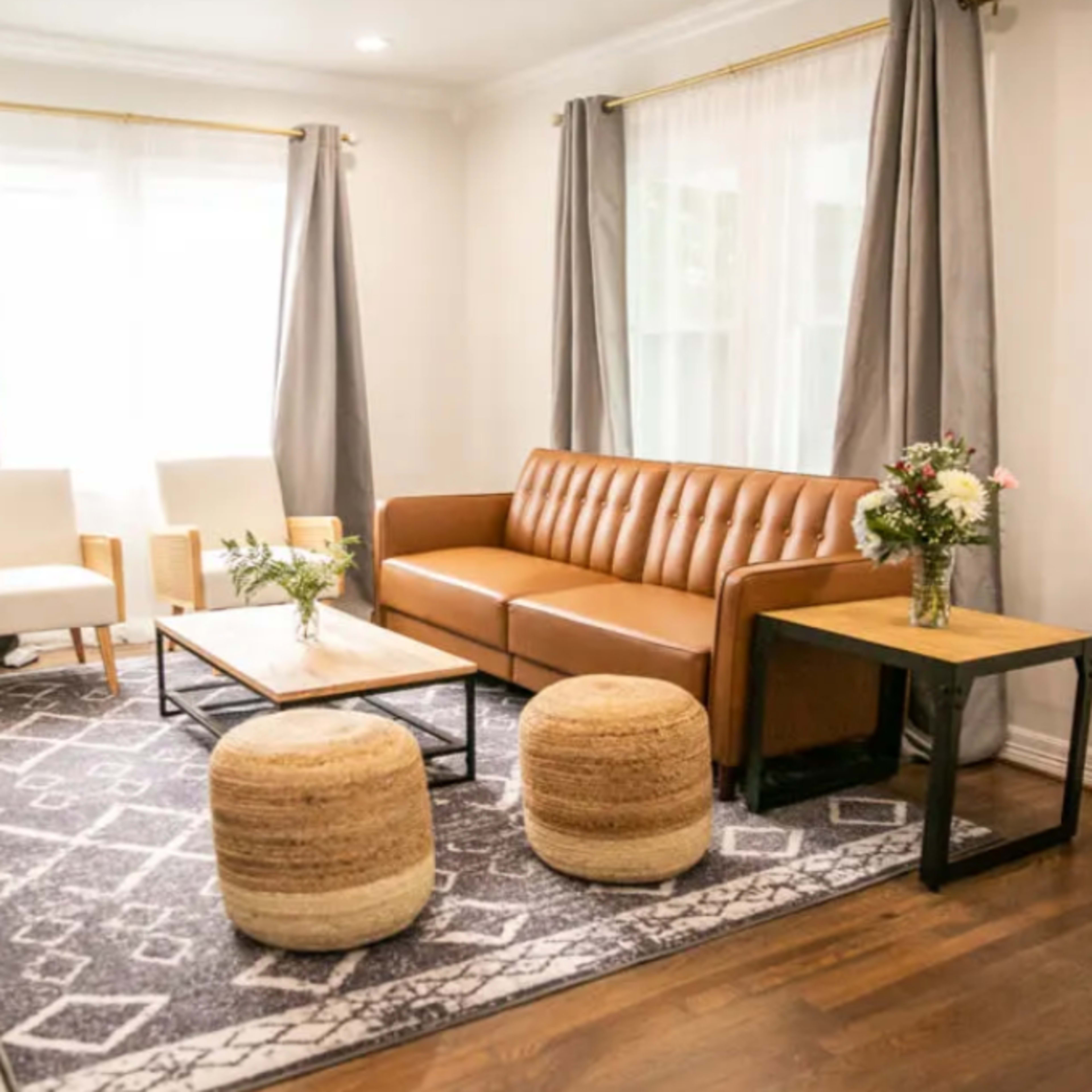 A modern living room with a brown leather sofa, two white chairs, a coffee table, and a woven area rug, surrounded by large windows with light gray curtains.