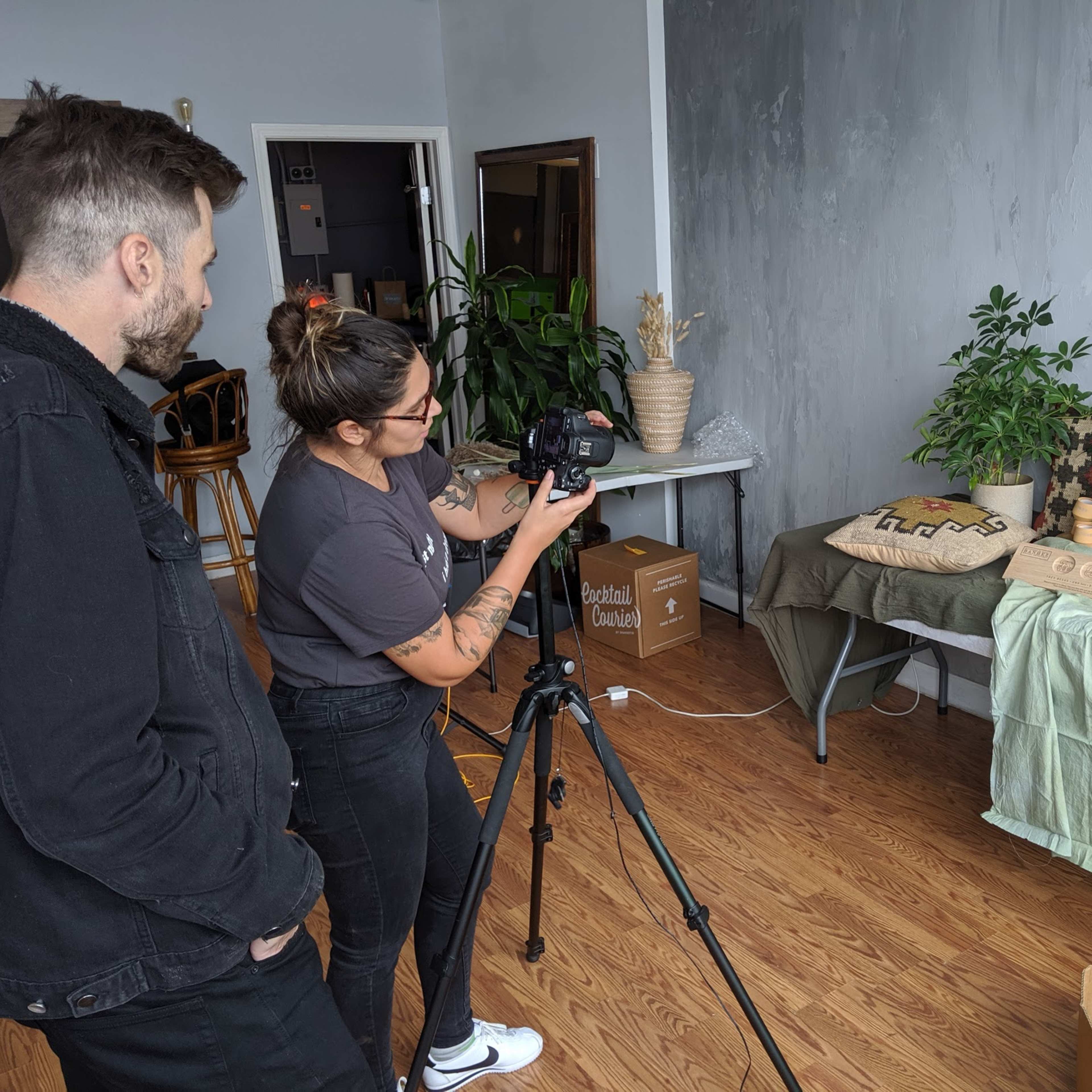 Two people are setting up a camera on a tripod to photograph a display of plants and products on a table in a well-lit room.