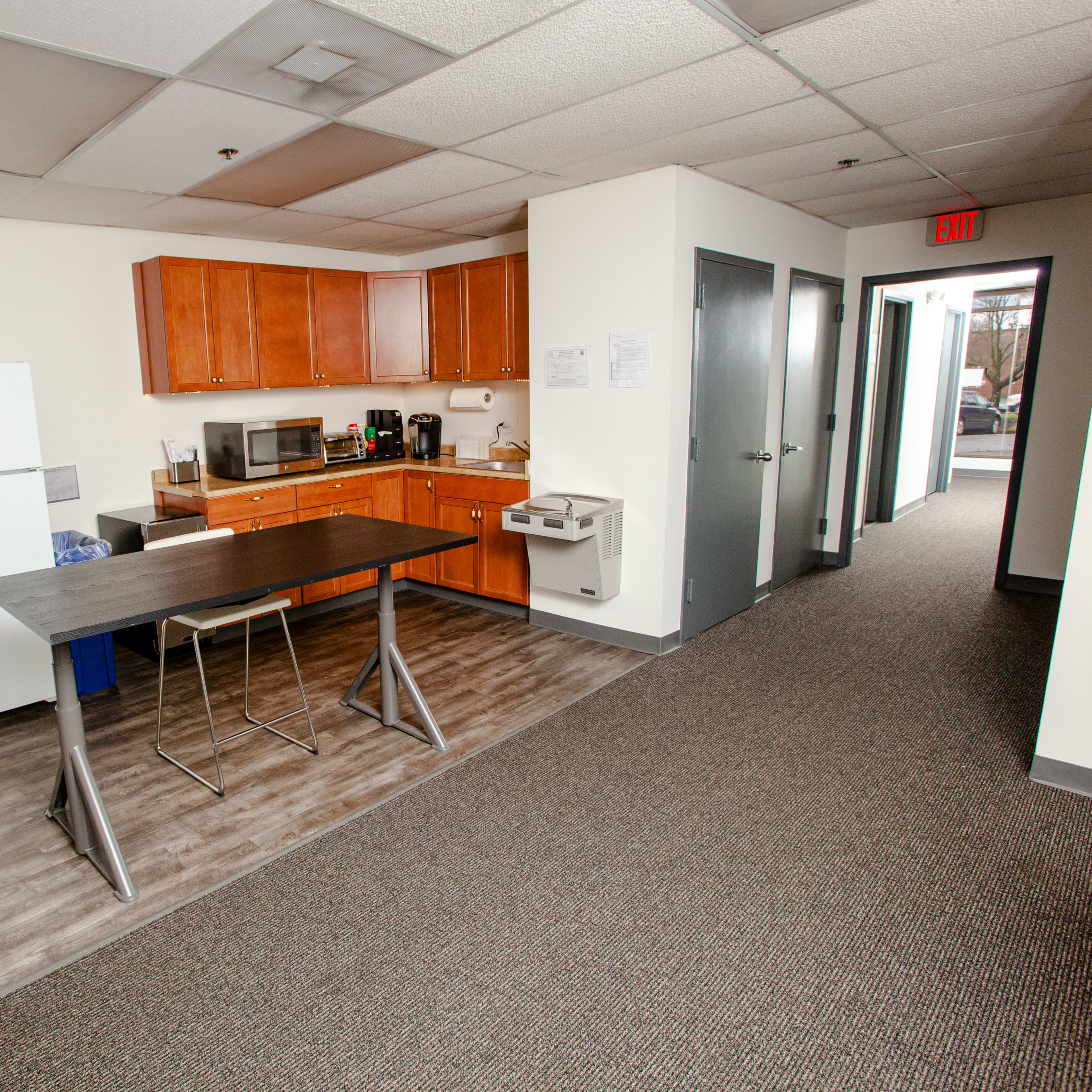 A kitchen area with wooden cabinets, a large refrigerator, a microwave, and a counter with two bar stools, adjacent to a hallway with carpeted flooring.