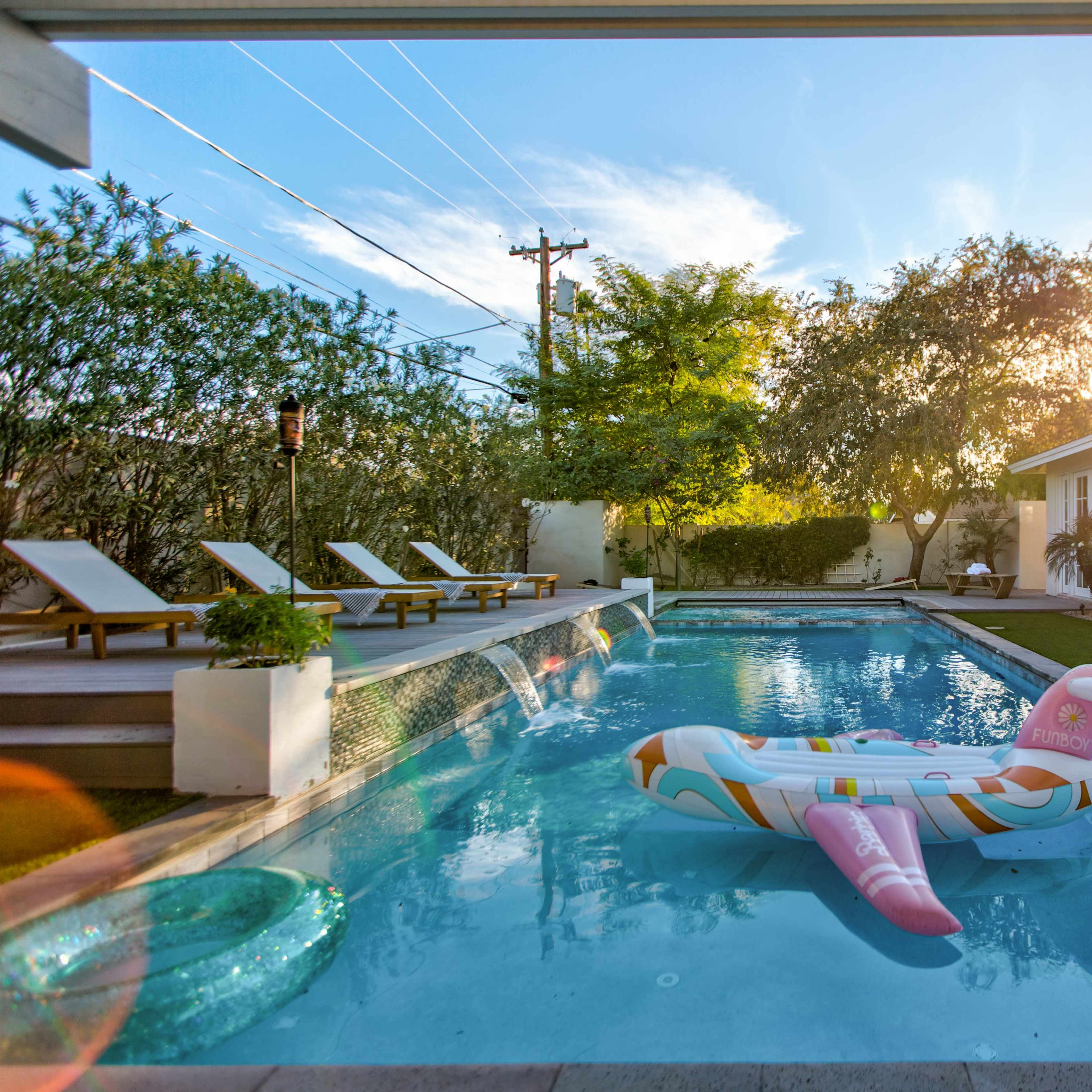 A swimming pool with a colorful inflatable float is surrounded by lounge chairs and greenery under a sunny sky.