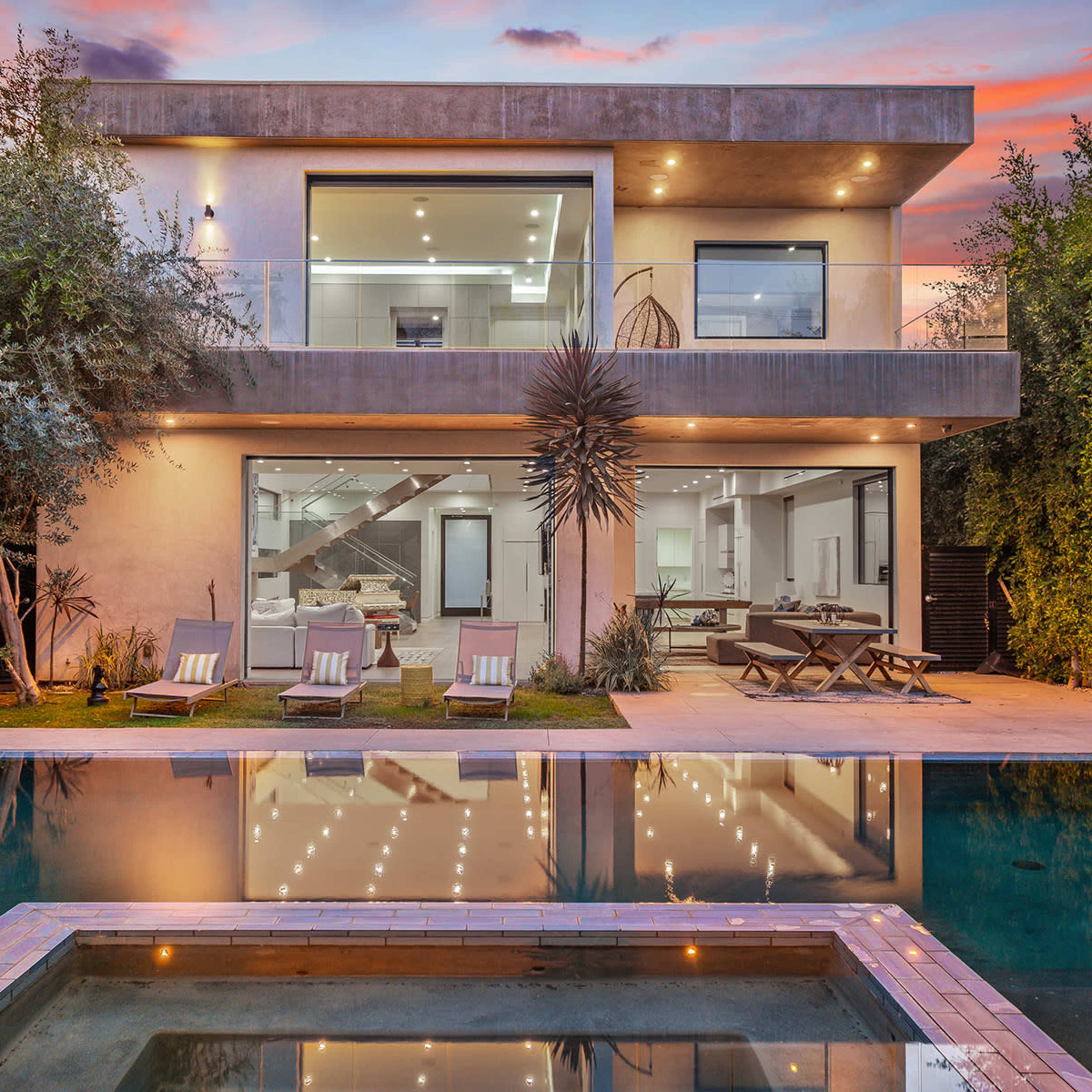 A modern two-story house with large windows is reflected in a swimming pool, surrounded by greenery and lounge chairs.