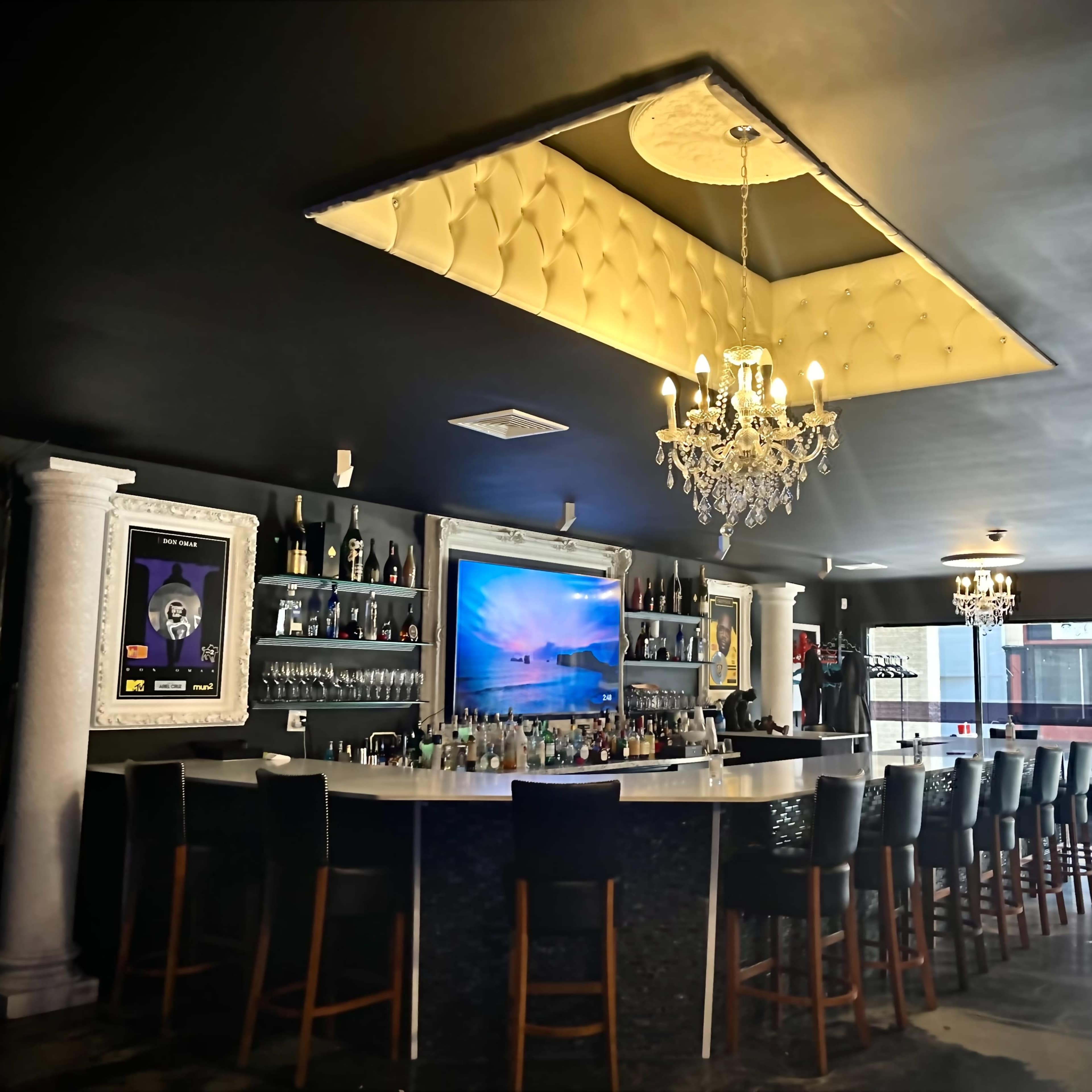 A modern bar with a curved white countertop, high-backed chairs, and a large screen displaying artwork, all set beneath a stylish chandelier.